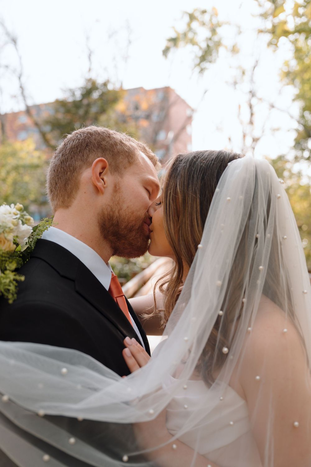 Romantic wedding couple sharing intimate kisses beneath a flowing veil against autumn foliage.