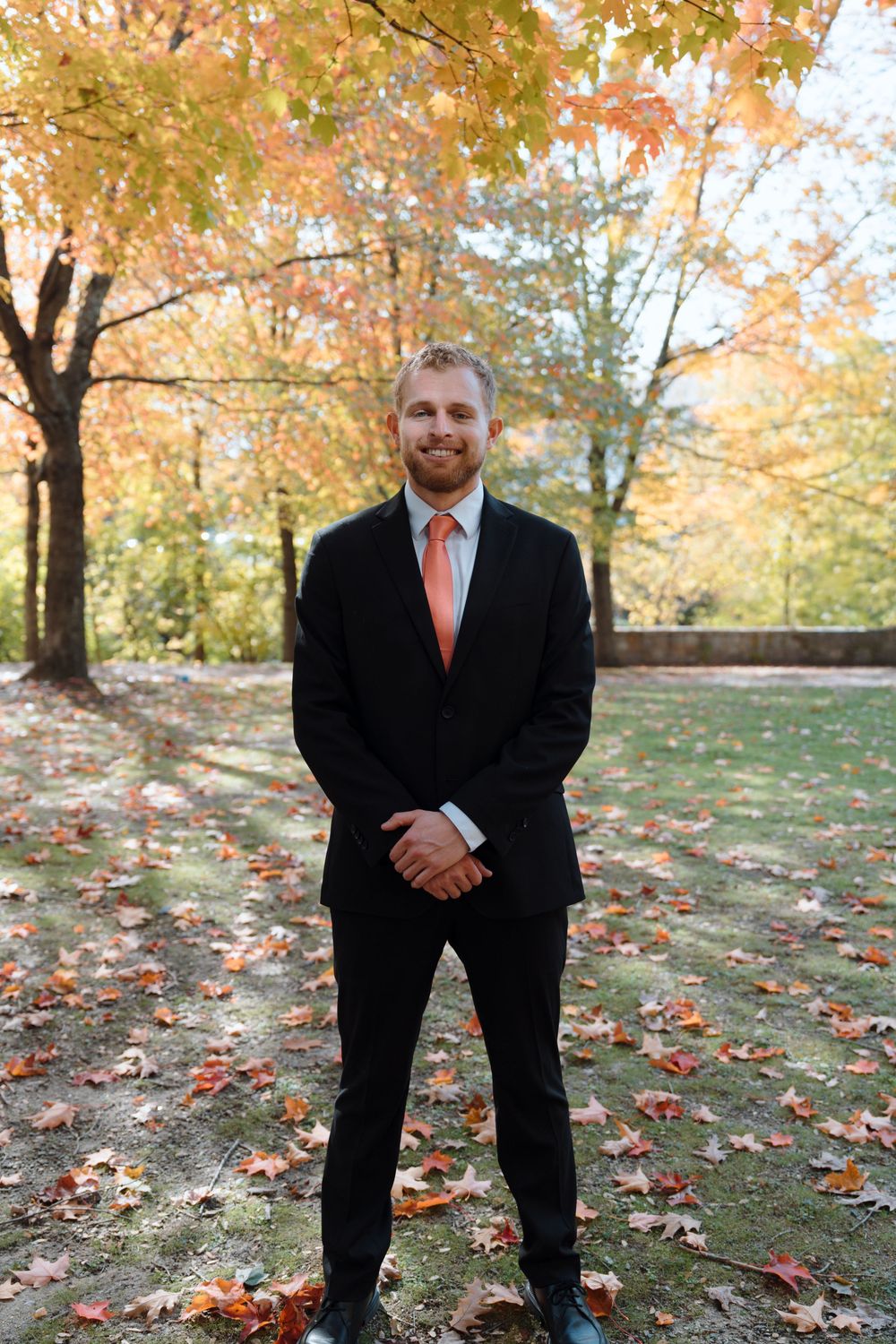 A person in a black suit with orange tie stands smiling in a fall setting with colorful autumn leaves.