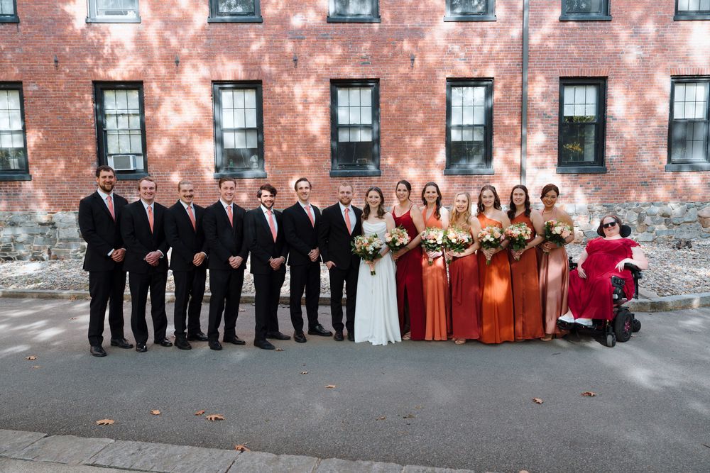 Wedding party in formal attire poses in front of rustic brick wall with bridesmaids wearing burnt orange dresses.