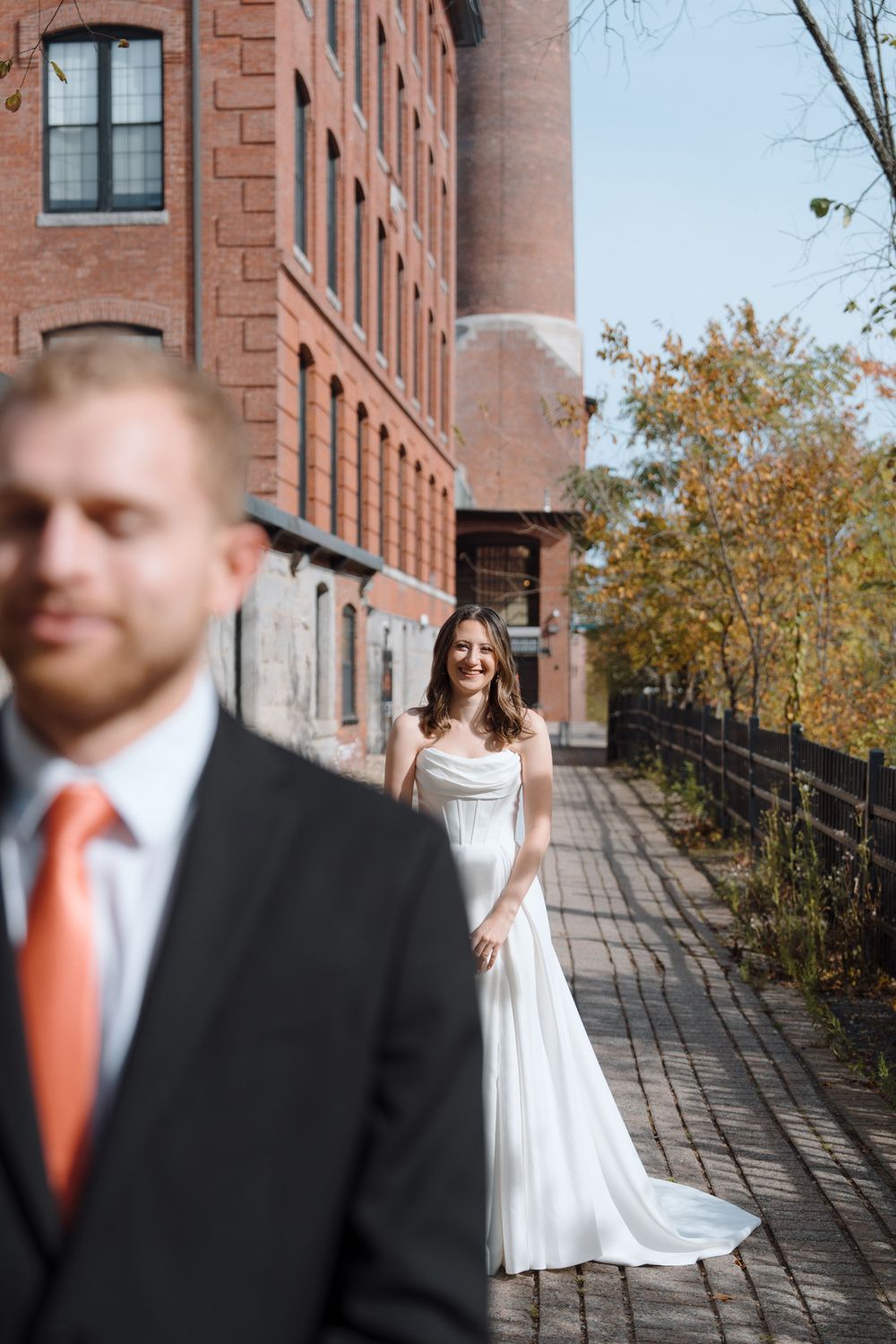A couple in wedding attire stands on a brick walkway outside historic buildings during an autumn day.