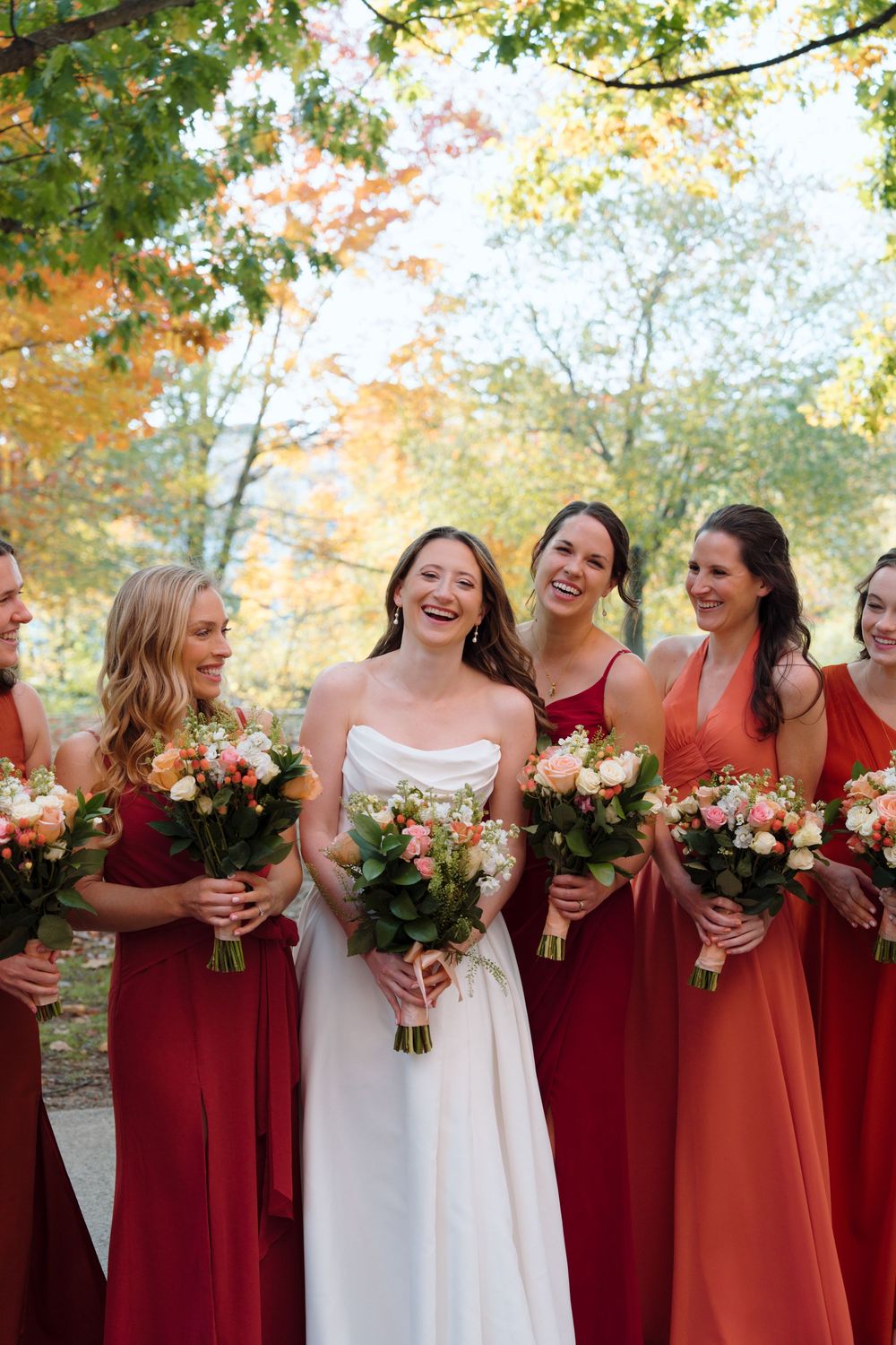 Wedding party in burgundy dresses holding bouquets against fall foliage background.