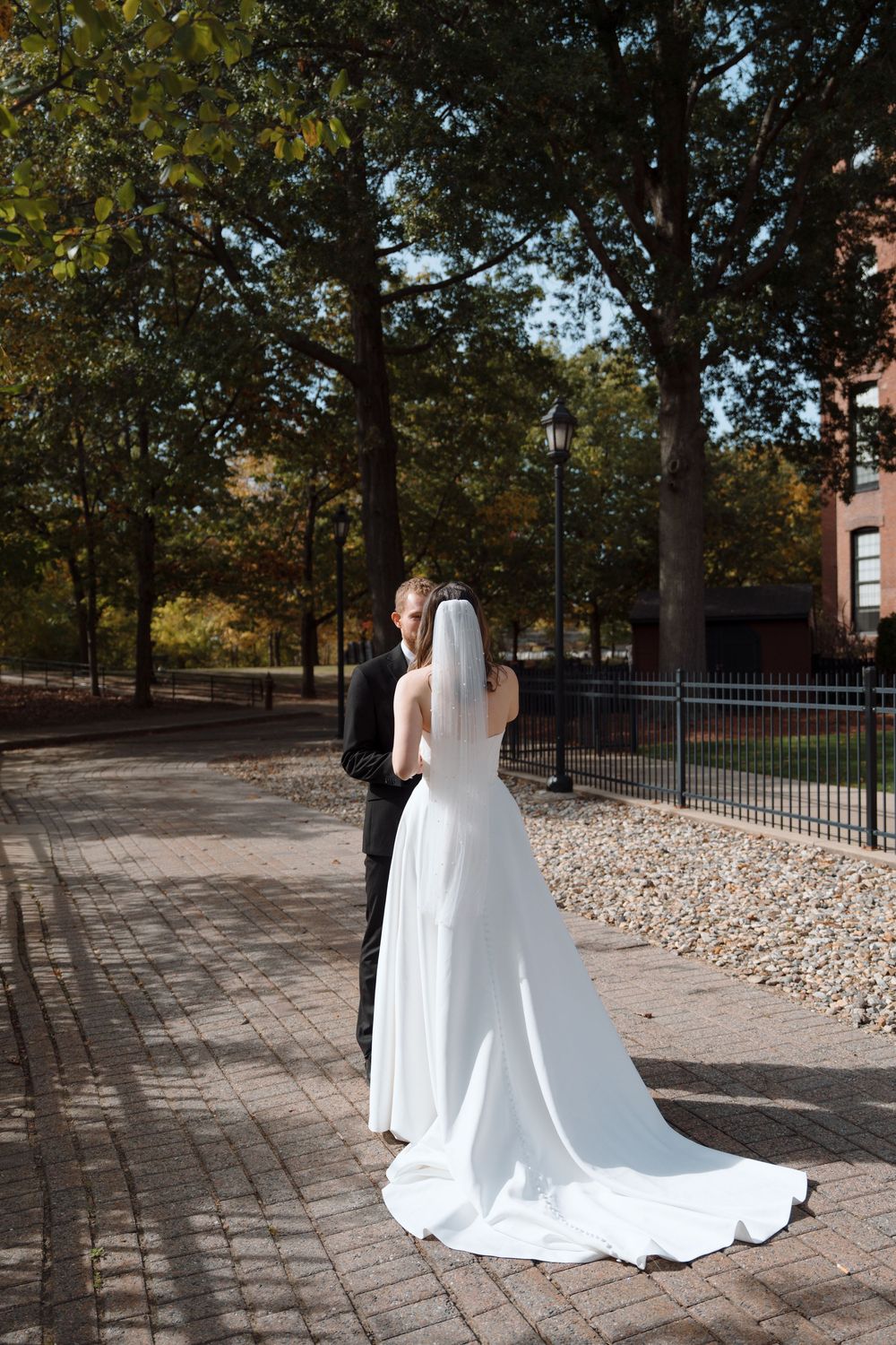 A wedding couple walks along a brick pathway surrounded by autumn trees in an outdoor garden setting.
