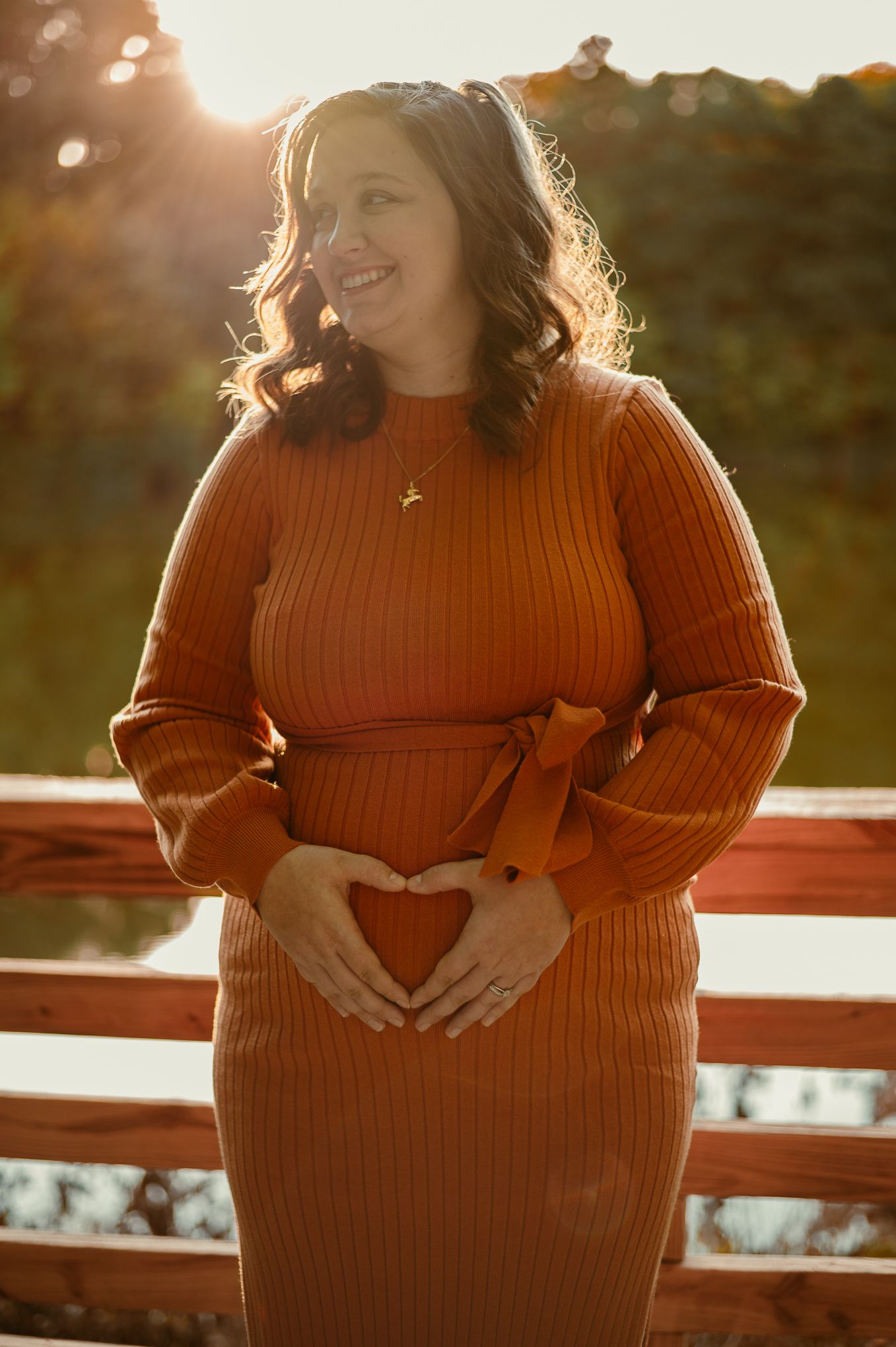 A maternity portrait of someone in a rust-colored sweater dress at sunset making a heart shape in Stafford VA.