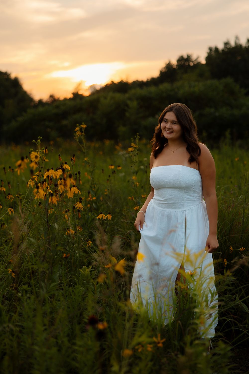 A woman in a white dress stands among blooming yellow wildflowers at sunset.