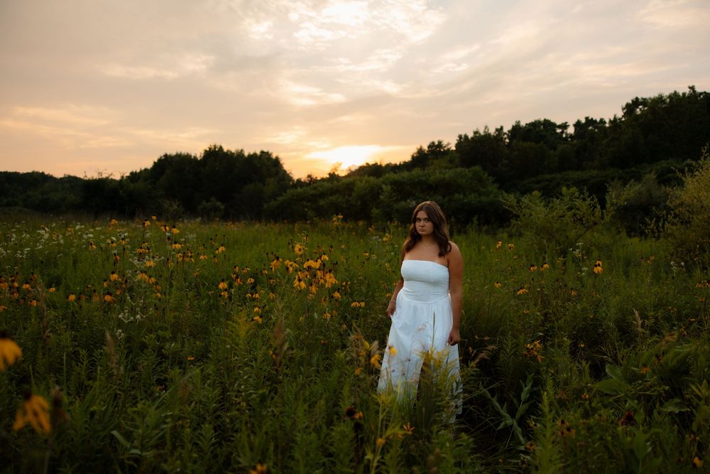 A person in a white dress stands in a field of wildflowers at sunset with warm golden light illuminating the scene.