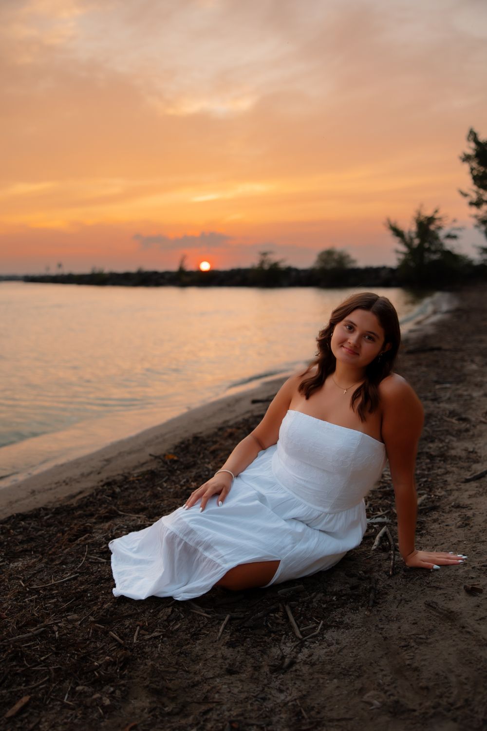 Beachside photo session at sunset with white dress on sandy shoreline near calm waters.