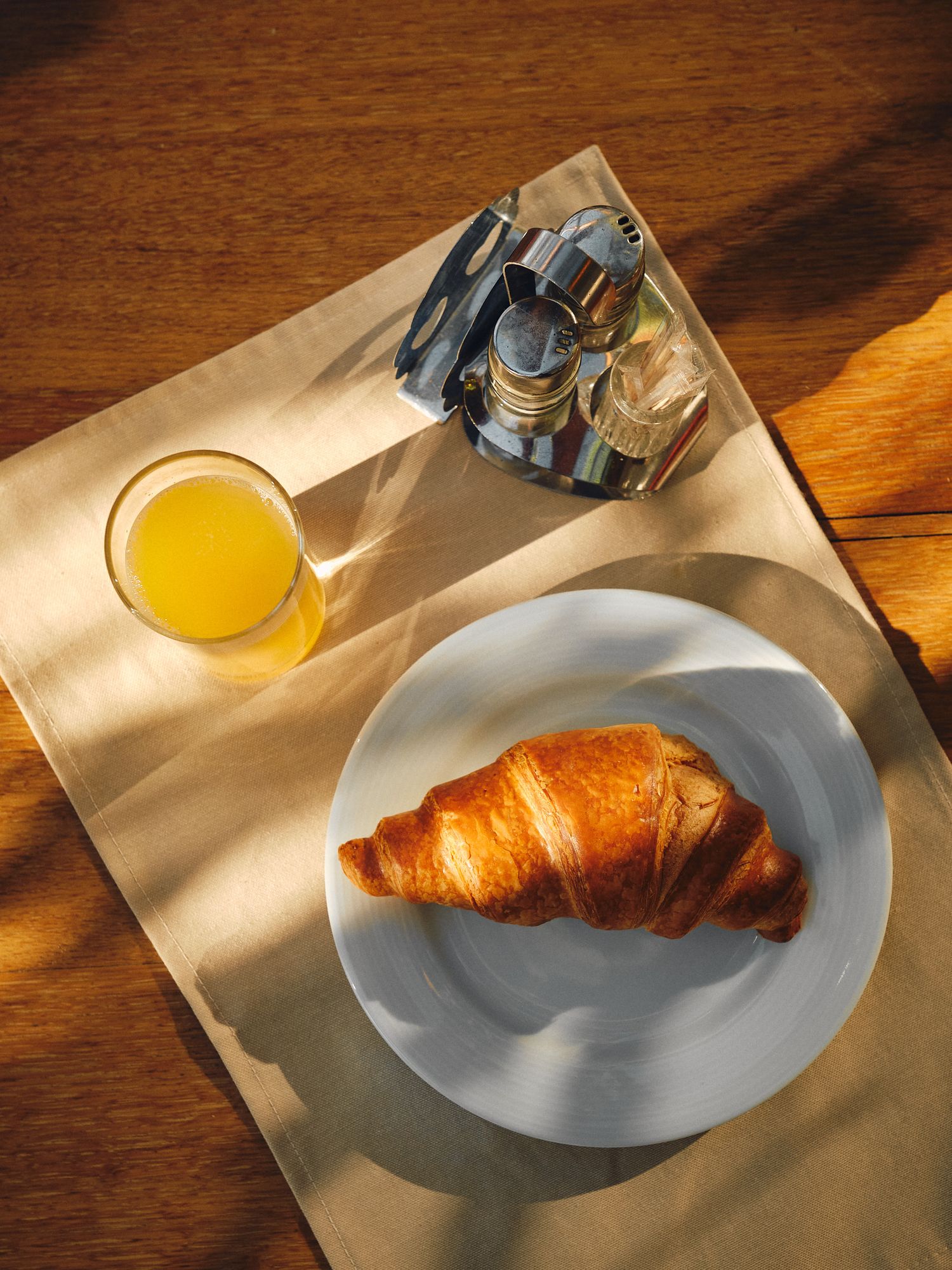 A cozy outdoor breakfast setting with a croissant, glass of beer, and wooden furniture on a stone patio in warm sunlight.