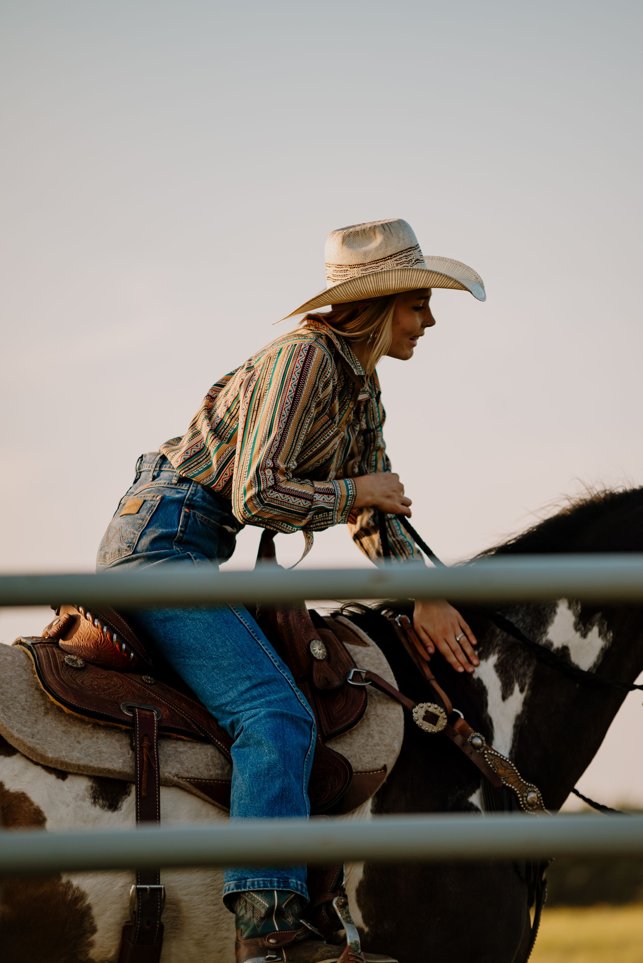 Cowboy in blue jeans and tan shirt leans forward while sitting on a fence during sunset.