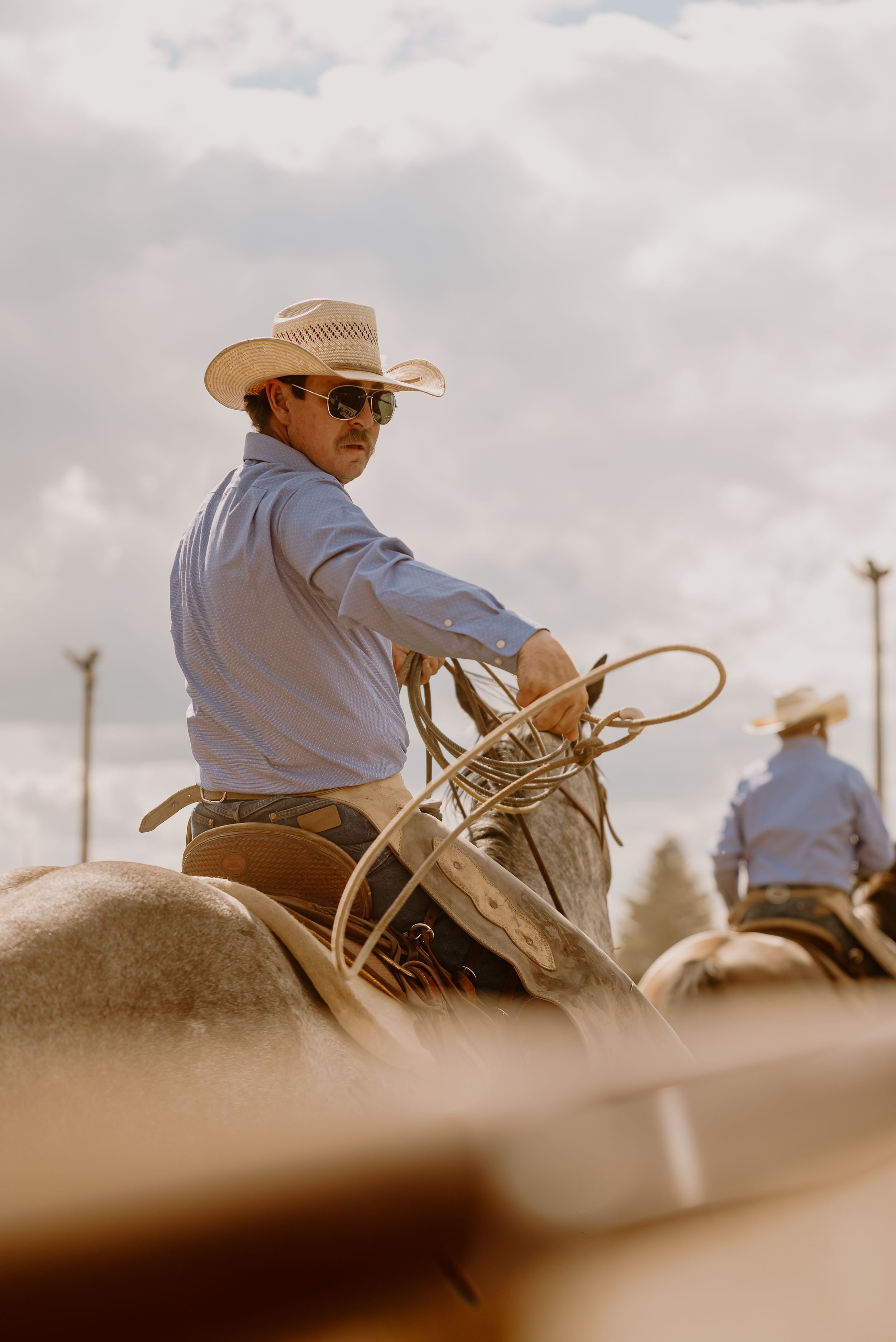 A cowboy wearing a light-colored hat and shirt rides horseback against a cloudy sky in a rustic western setting.