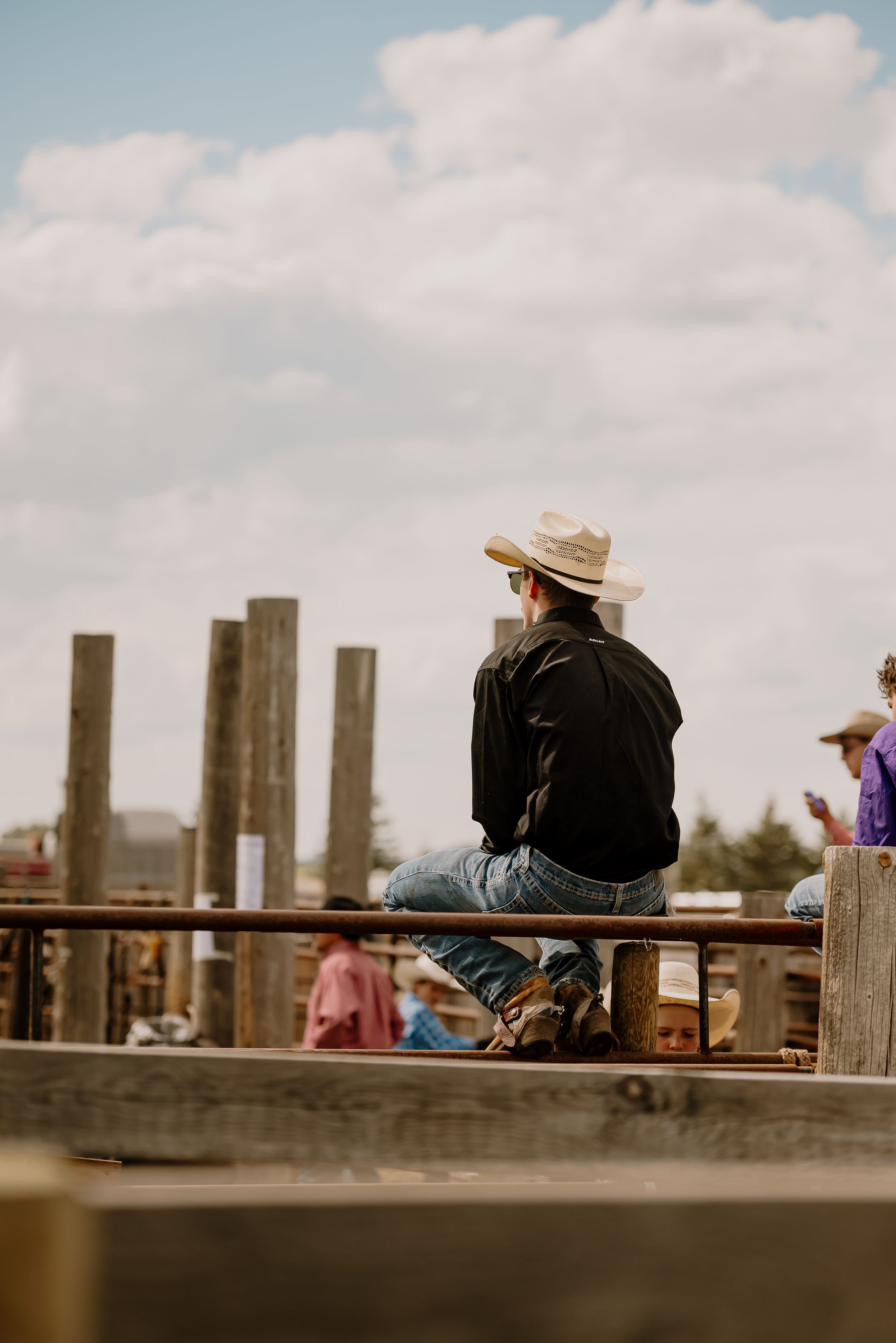 People in cowboy hats sit on a wooden pier railing overlooking a scenic view on a cloudy day.