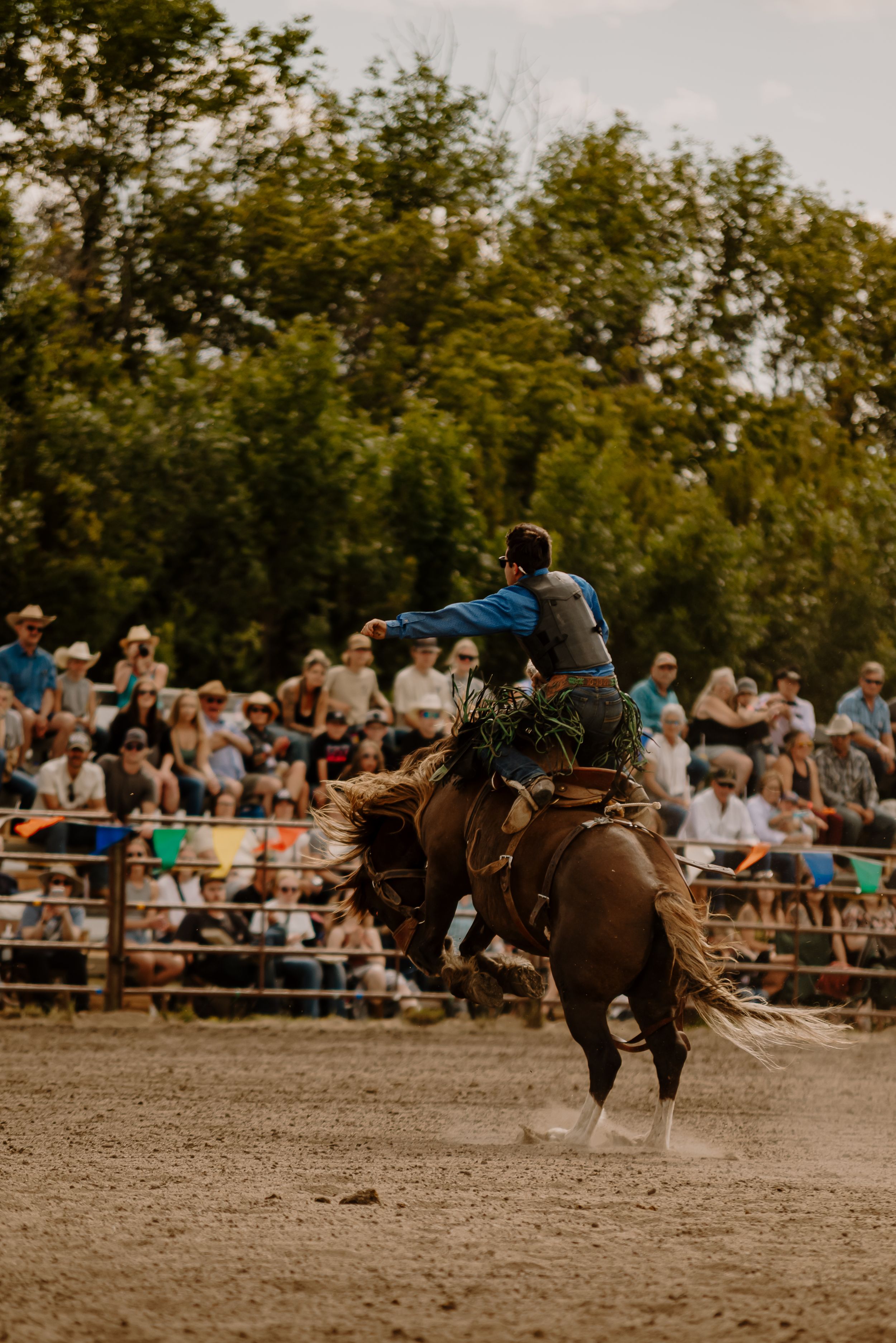 A rodeo rider competes in a bronc riding event at a dusty arena with spectators watching from the stands.