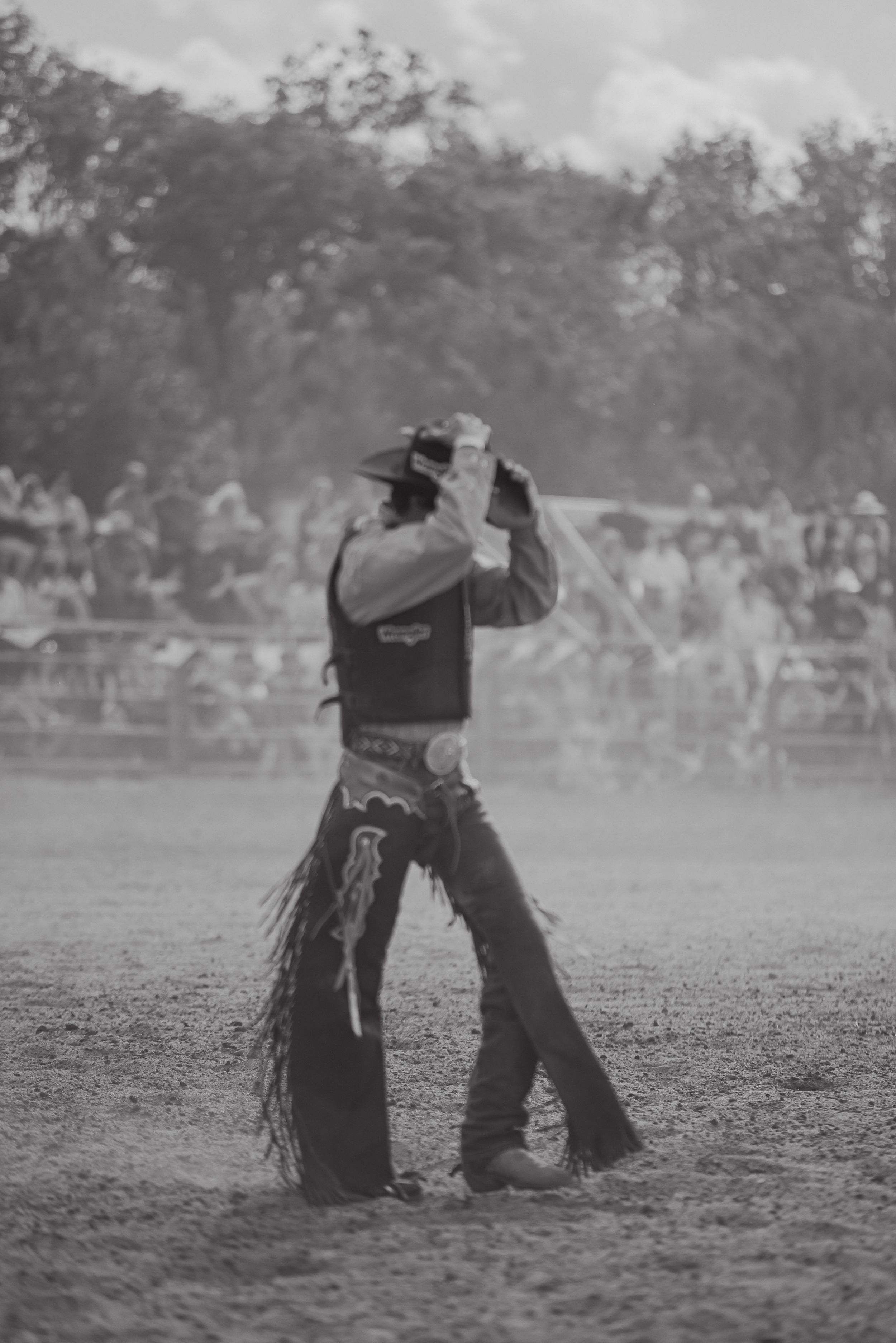 A rodeo performer in traditional western chaps and hat stands in a dusty arena with spectators in the background.