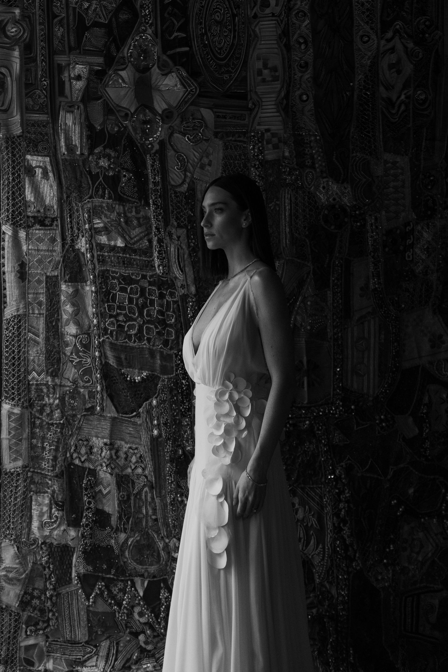 A dramatic black and white portrait of a figure in a white dress against an ornate patterned wall.