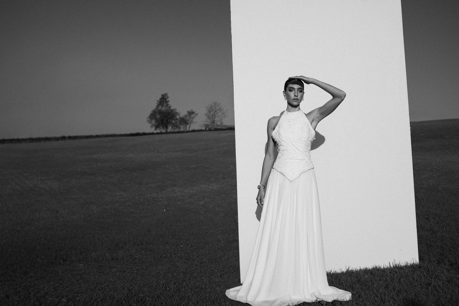 A figure in a flowing white dress poses dramatically against a stark white backdrop in an outdoor setting at dusk.