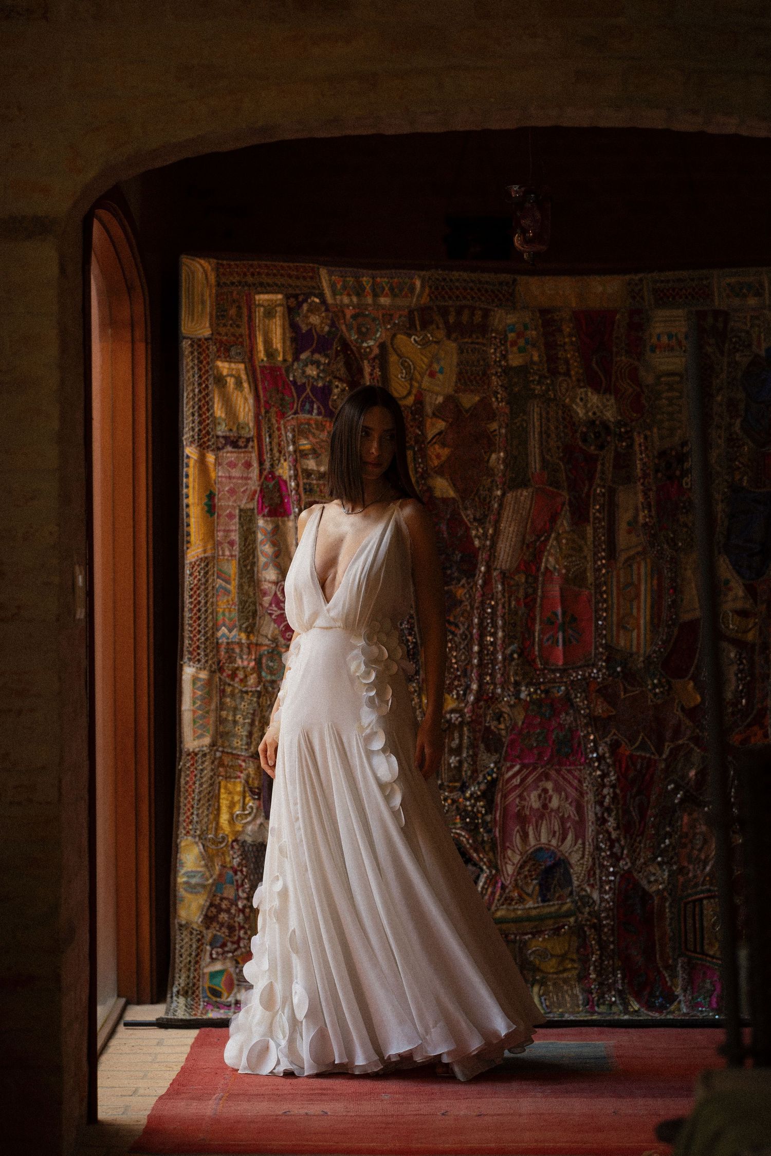 A flowing white wedding dress photographed against an ornate tapestry backdrop in a dimly lit room.