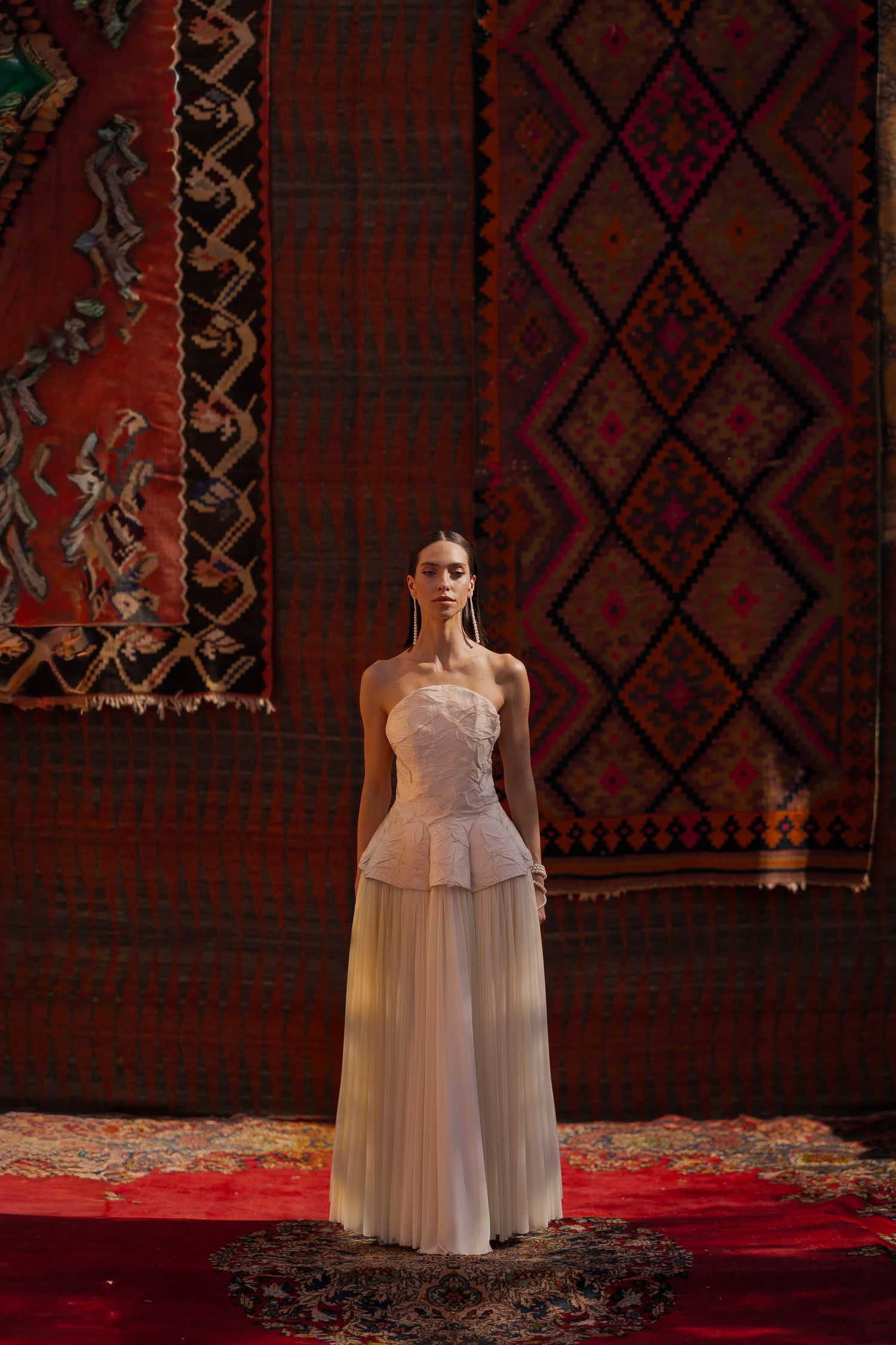 A figure in a flowing white dress stands against an ornate patterned backdrop with red carpet.