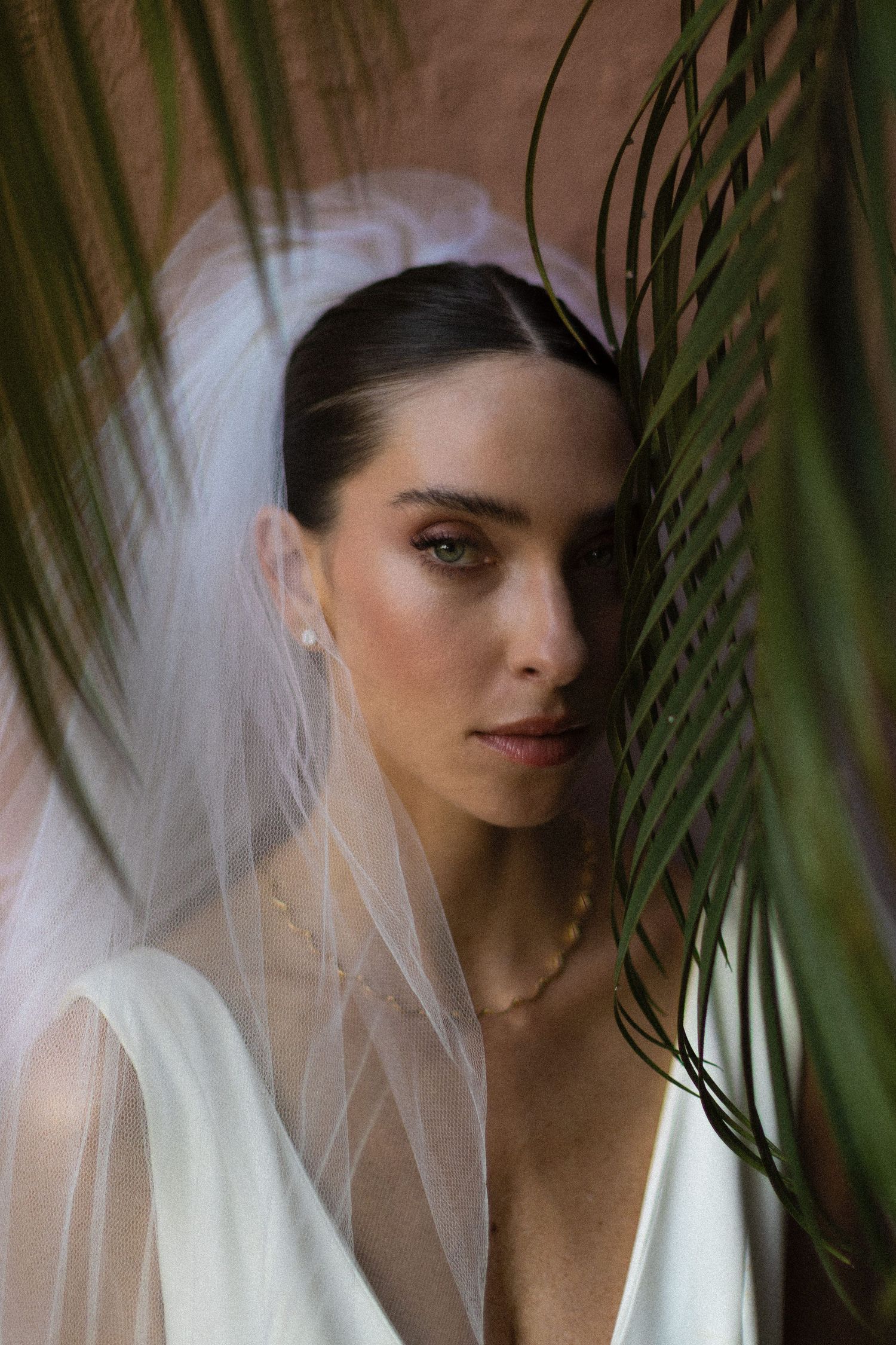 Artistic bridal portraits through palm leaves showing a white veil and elegant makeup.