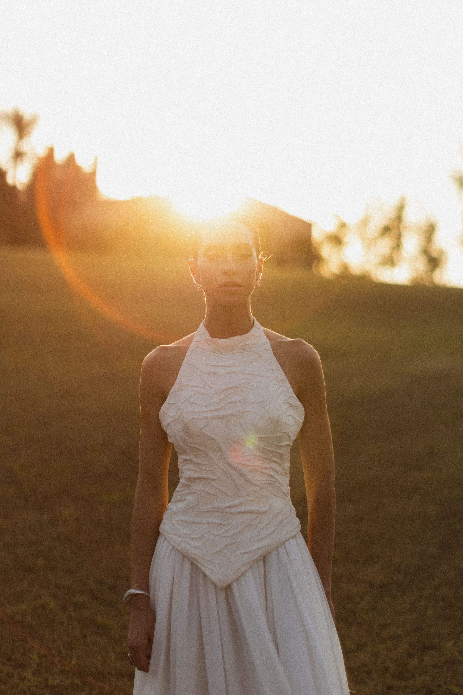Figure in white dress stands silhouetted against golden sunset light in field.