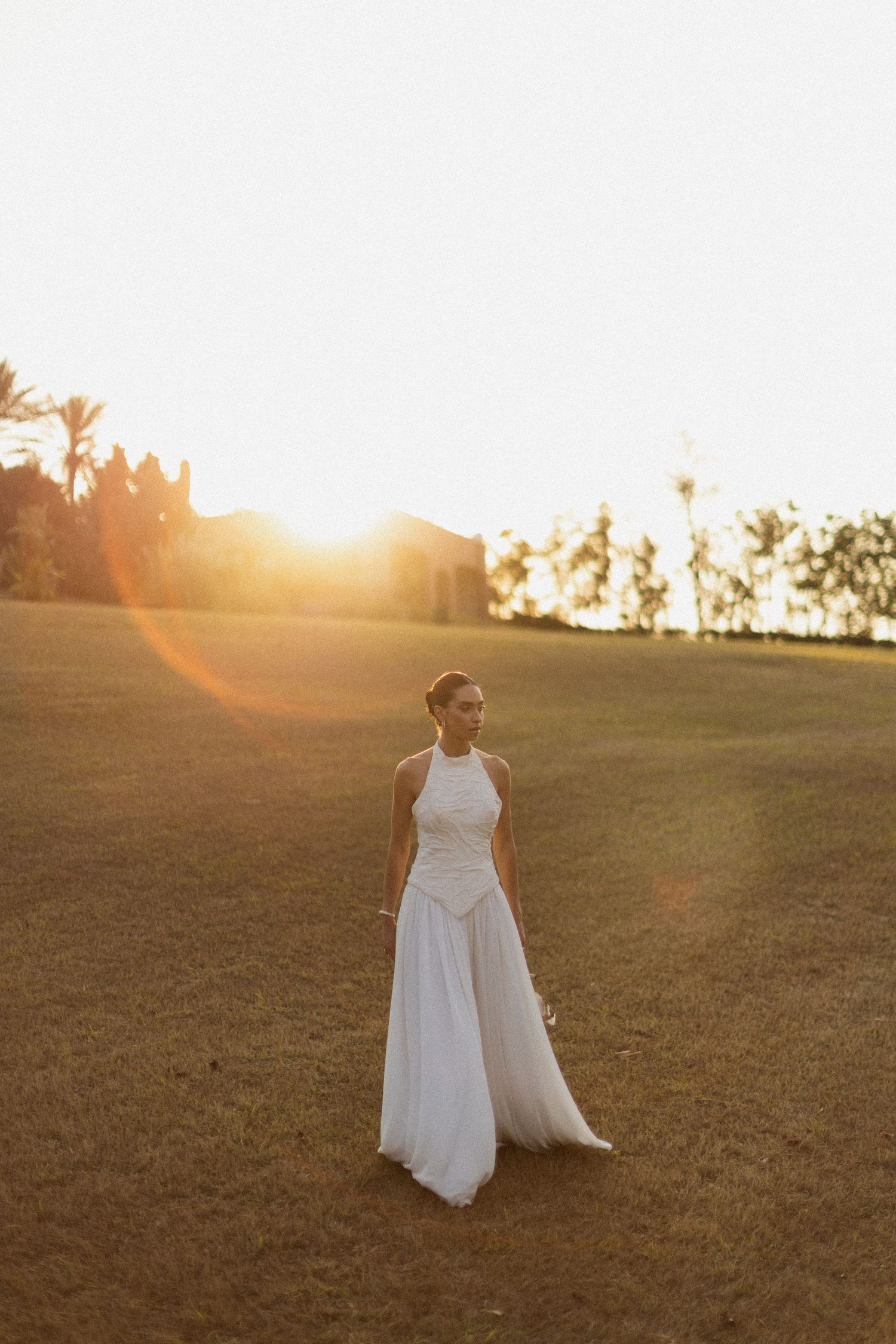 Figure in flowing white dress stands in grassy field during golden sunset with palm trees in background.