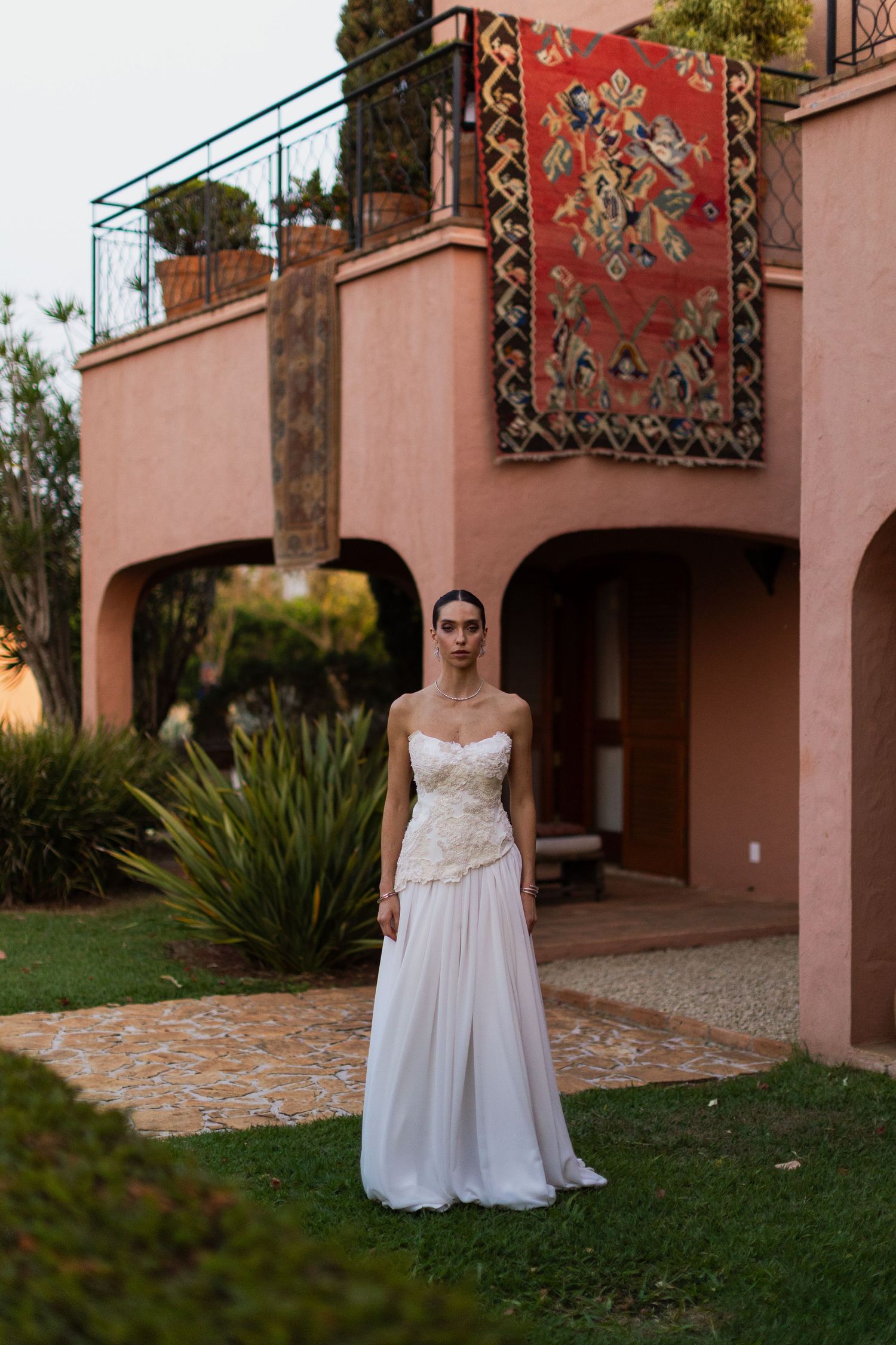 Elegant white wedding gown against pink Mediterranean-style villa with decorative carpet hanging on balcony.