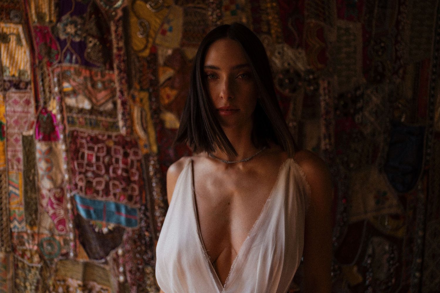 Artistic portrait in dim lighting with colorful patterned tapestries in background.