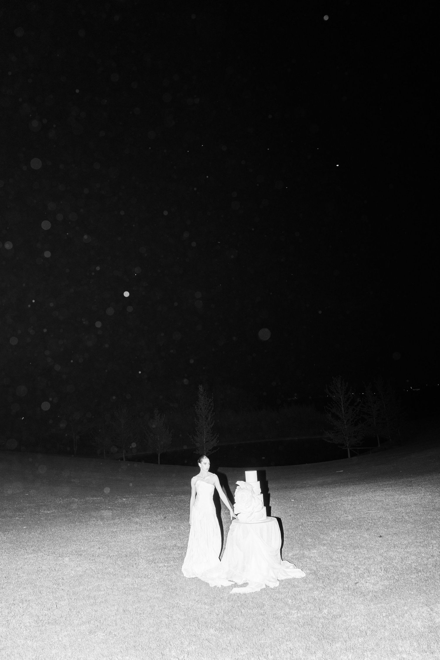Black and white nighttime photo of a wedding dress and figure in an open field under starry sky.