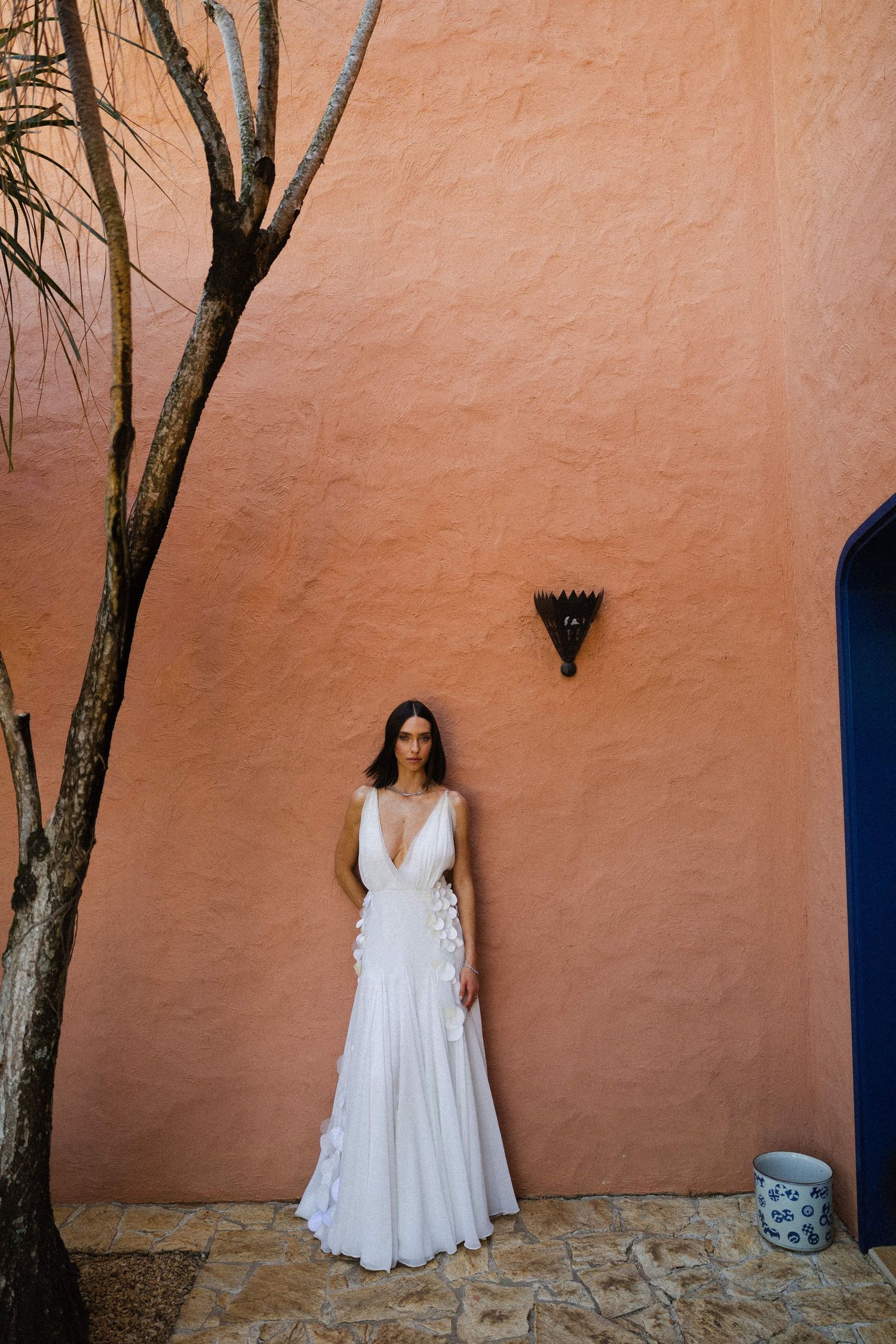 White wedding dress photographed against coral-colored stucco wall with blue archway and tree.