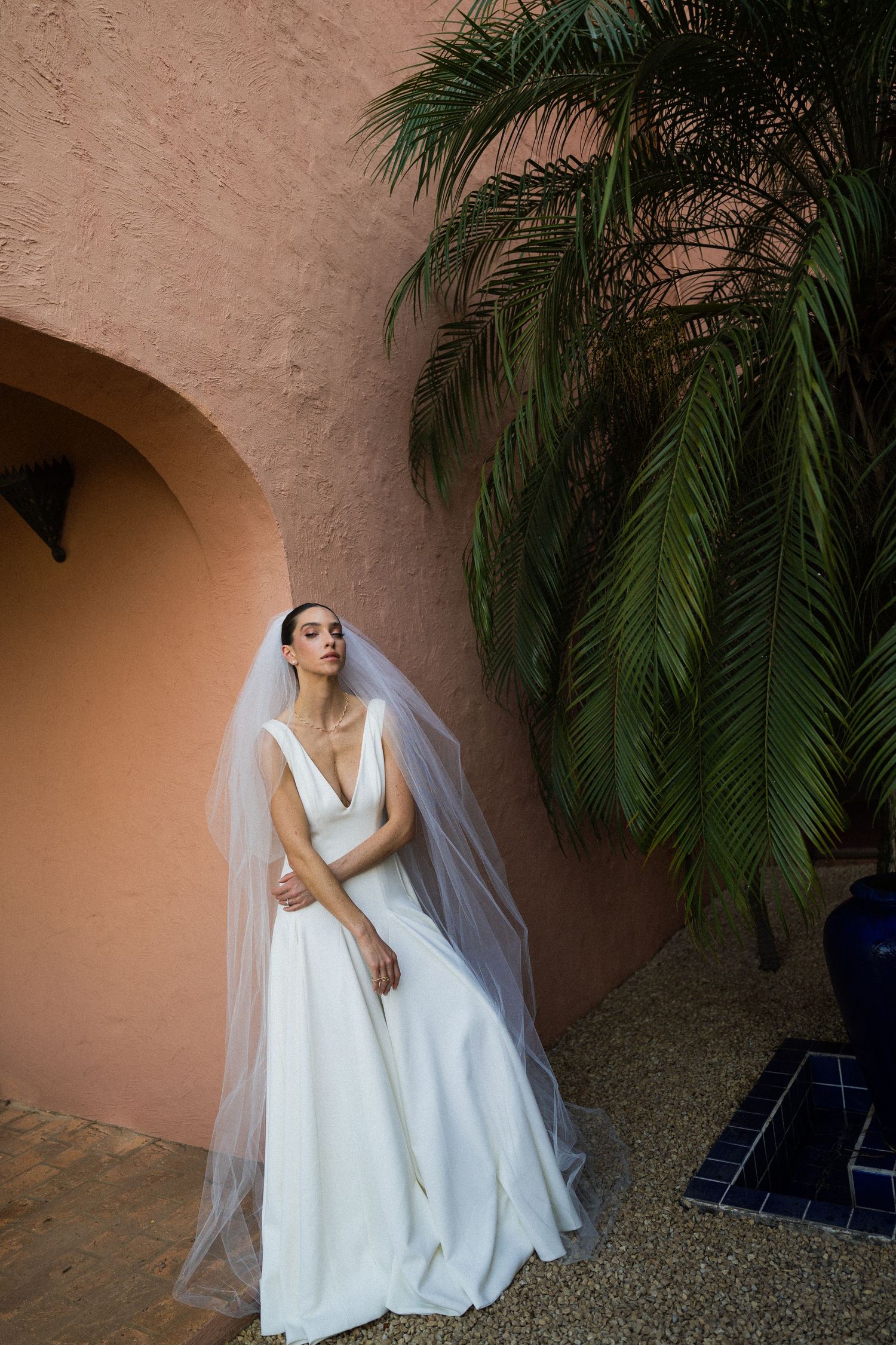 A bride in an elegant white wedding dress and veil poses against a textured wall with tropical palm fronds.