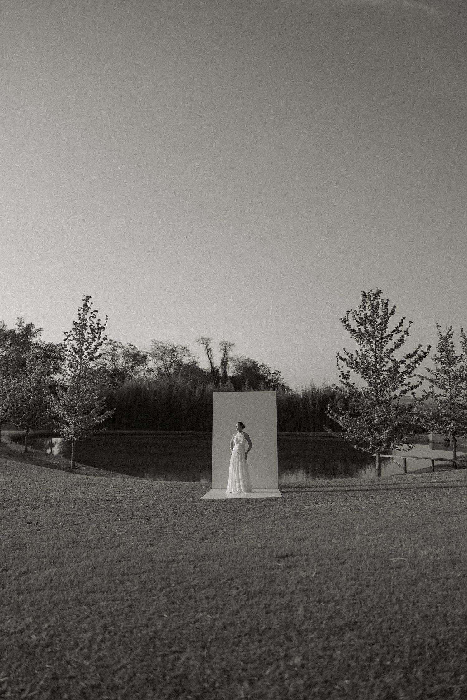 Black and white artistic photo of a figure in white standing against a monolithic panel outdoors near a lake and trees.