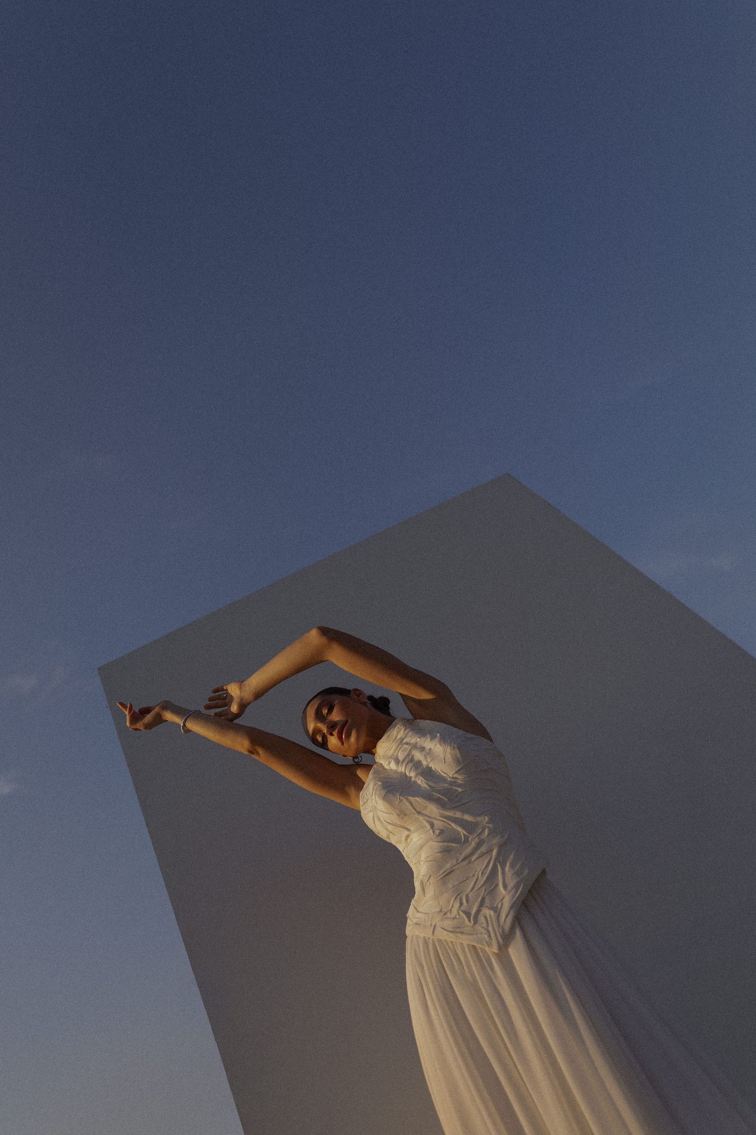A person in a white dress poses gracefully against a dark blue sky at dusk.