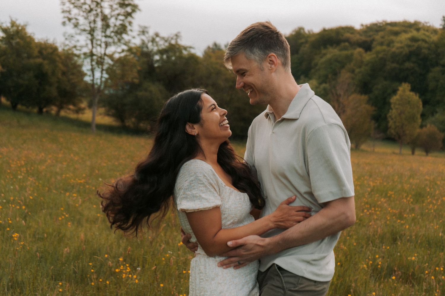A happy couple embraces and laughs together in a sunny wildflower meadow with hills behind them.