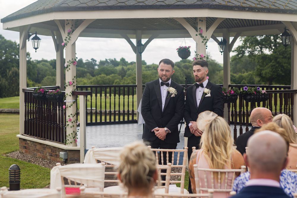 Wedding guests seated at an outdoor ceremony venue with white chairs and gazebo.