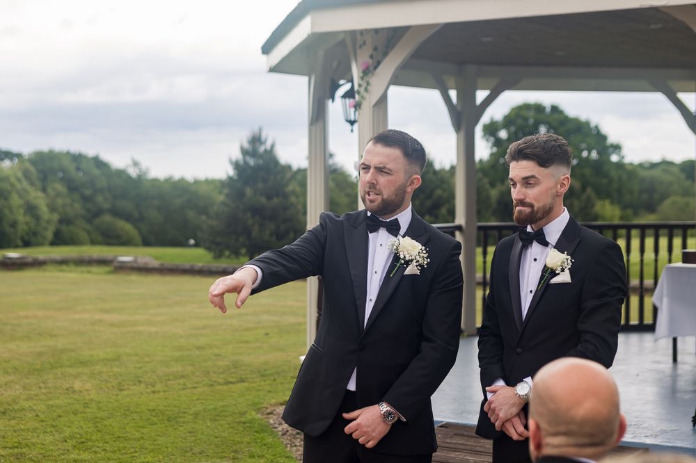 Two groomsmen in black tuxedos stand under a white gazebo during an outdoor wedding ceremony.