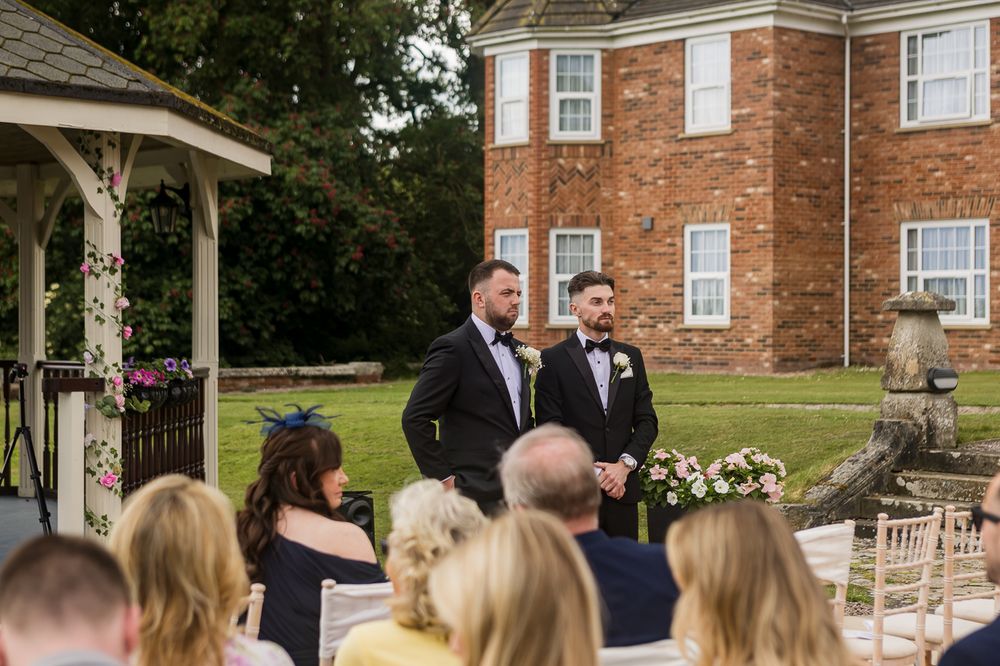 Wedding ceremony takes place under a gazebo with guests seated on white chairs in front of brick building.