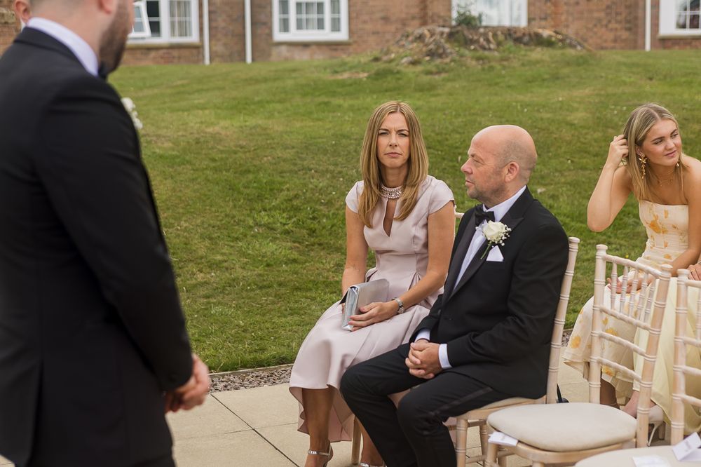 Wedding guests seated on white chairs during an outdoor ceremony near a brick building.