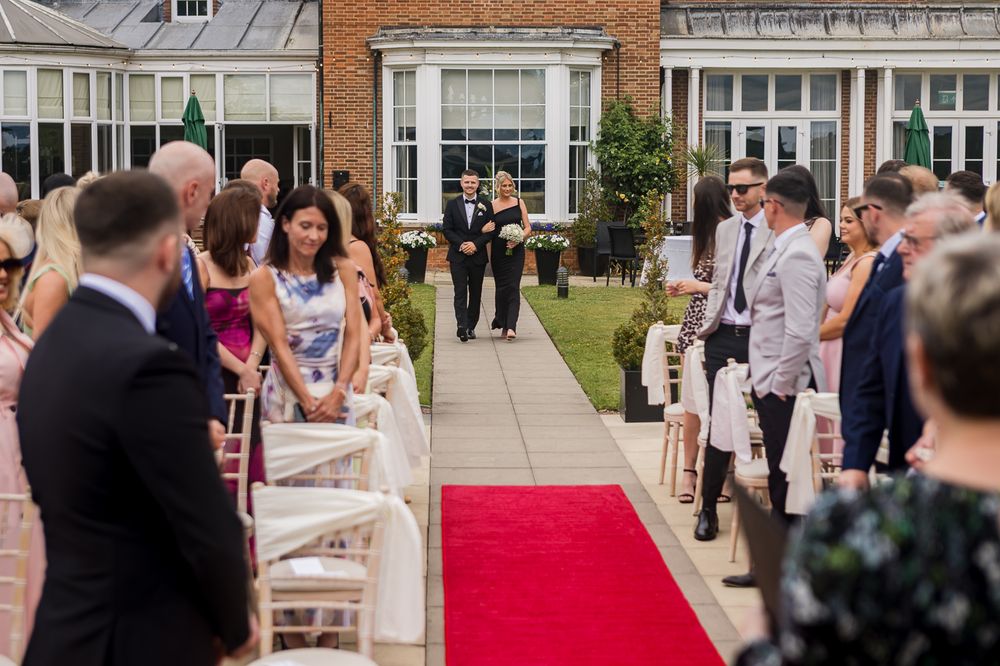 Wedding guests gather along a red carpet aisle outside a brick mansion for an outdoor ceremony.