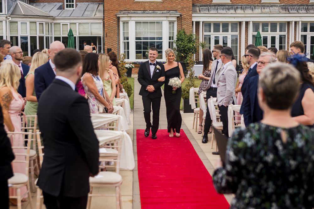 Wedding ceremony with red carpet aisle and guests seated on white chairs outside brick venue.