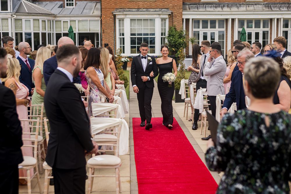 Wedding ceremony processional with guests lining red carpet aisle outside brick venue.