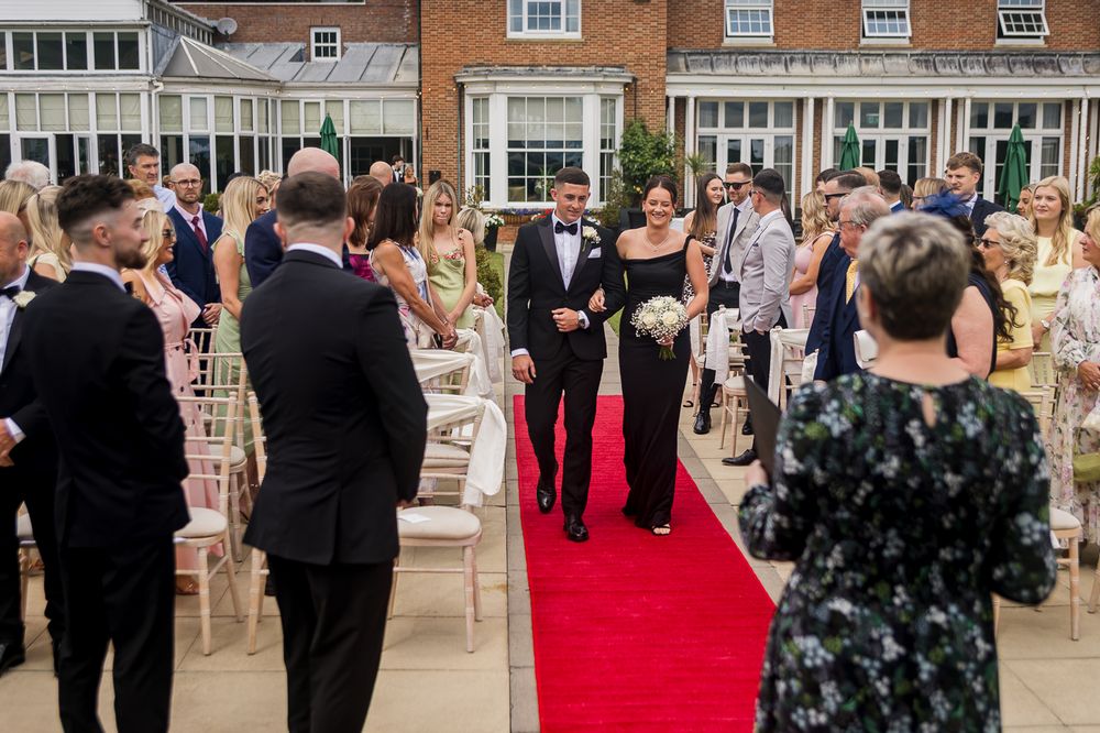 Wedding guests applaud during processional on red carpet at elegant outdoor ceremony.
