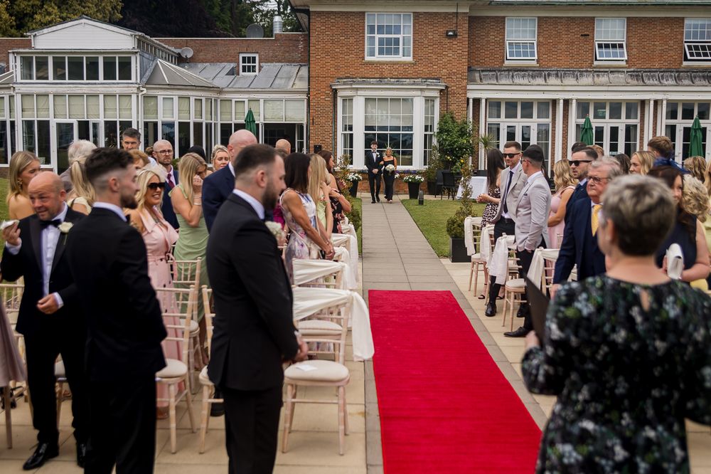 Outdoor wedding ceremony setup with red carpet aisle and white chairs against brick building backdrop.