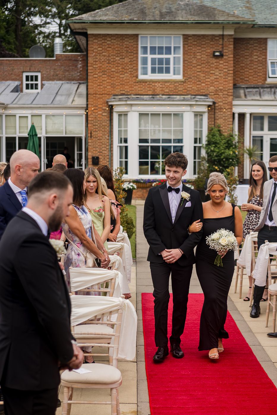 Wedding processional on red carpet with formal attendees in black tie attire.