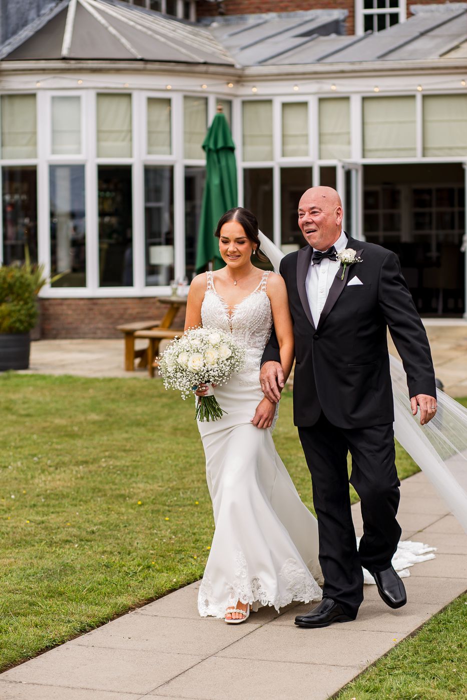 Wedding couple walks together on concrete path outside brick venue building.
