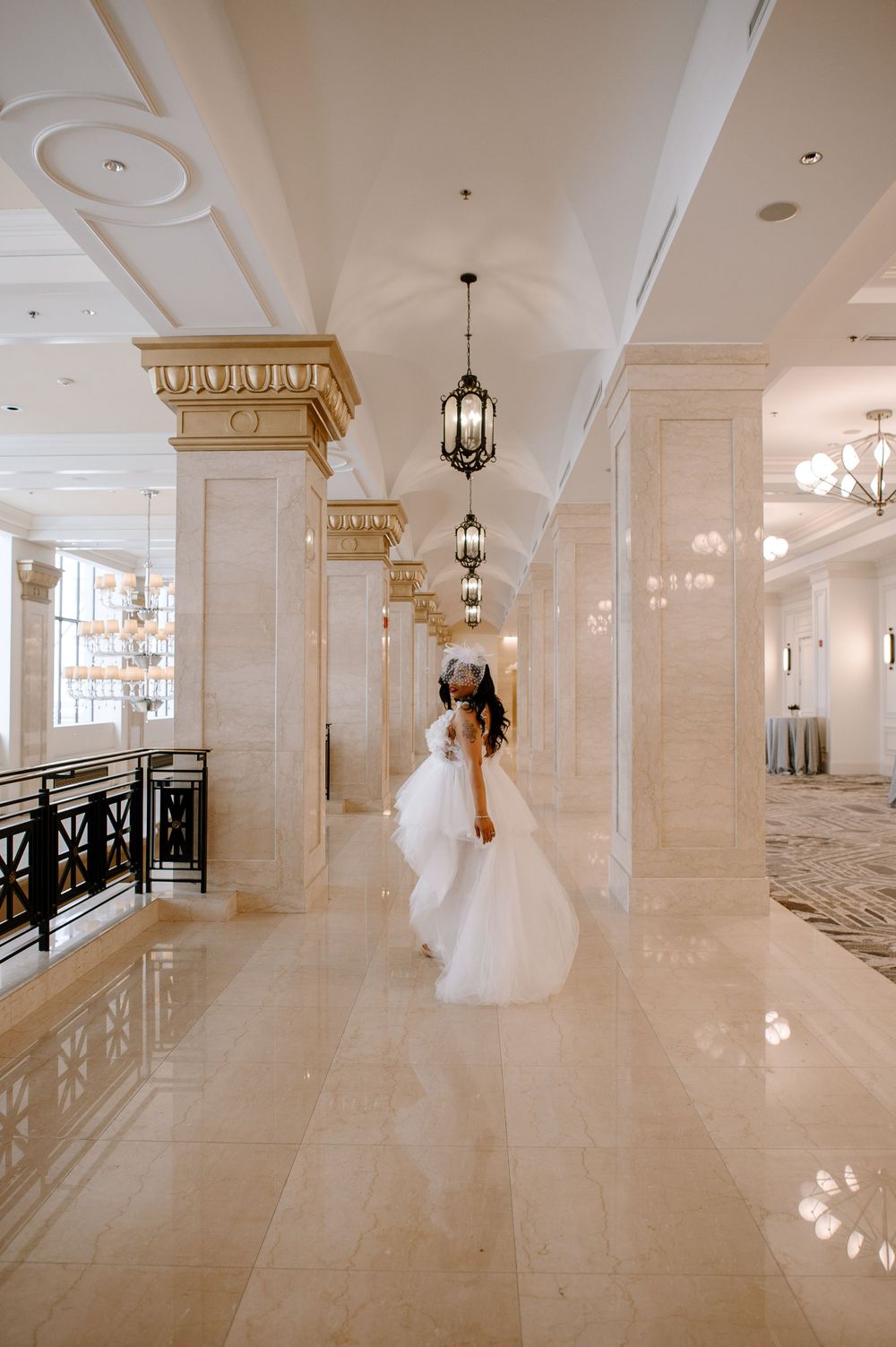 Elegant white hallway with ornate columns, chandeliers, and black railings in a luxury wedding venue.