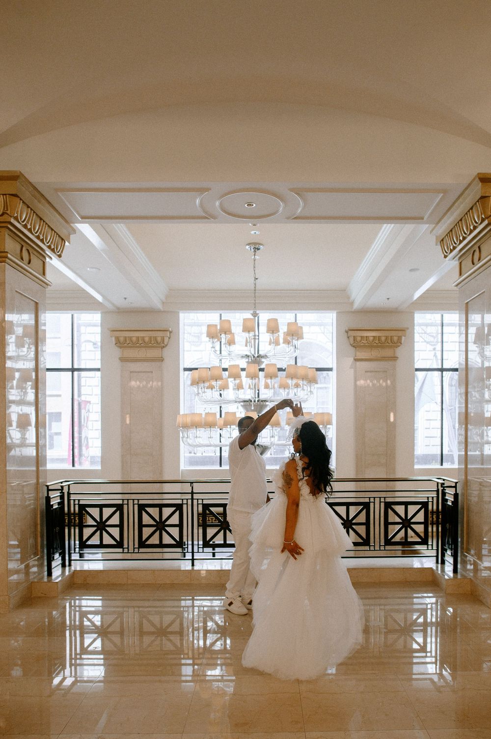 Wedding party members in white attire dance and pose in an elegant ballroom with ornate columns and chandeliers.