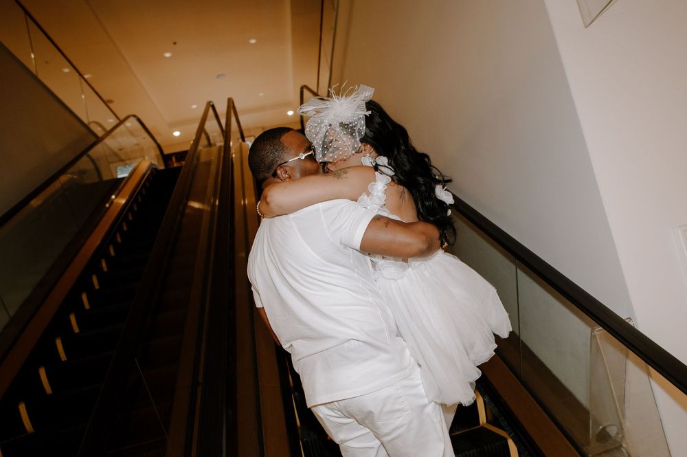 Couple in white attire walking up elegant staircase with black railings in romantic indoor setting.