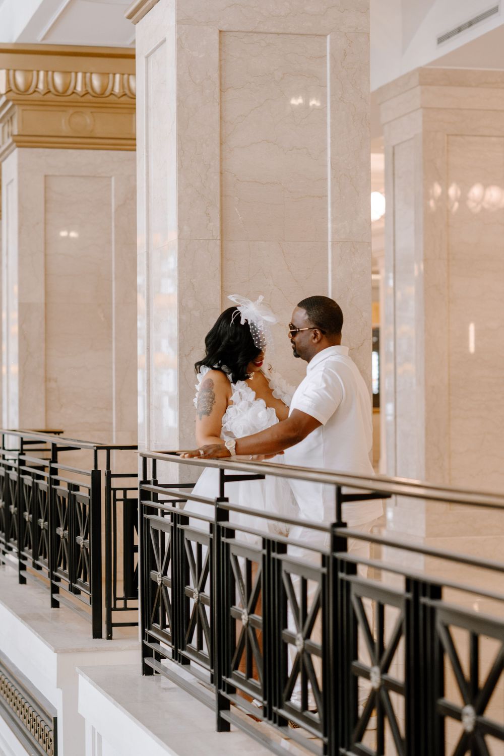 Bride in flowing white dress walks along a grand hallway with black decorative railings and cream walls.