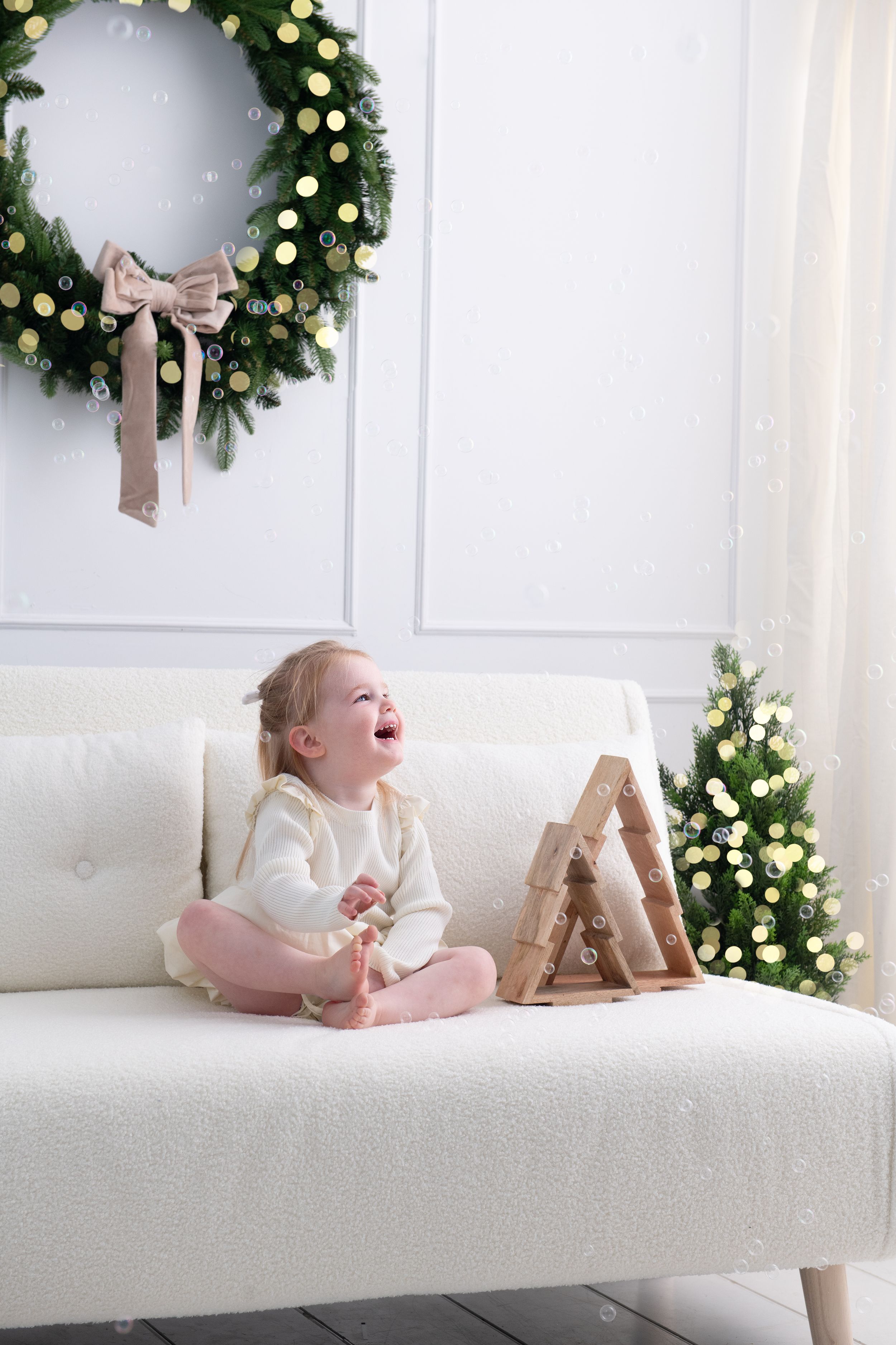 Baby in white dress sits on couch with wooden letters and Christmas wreath in background.