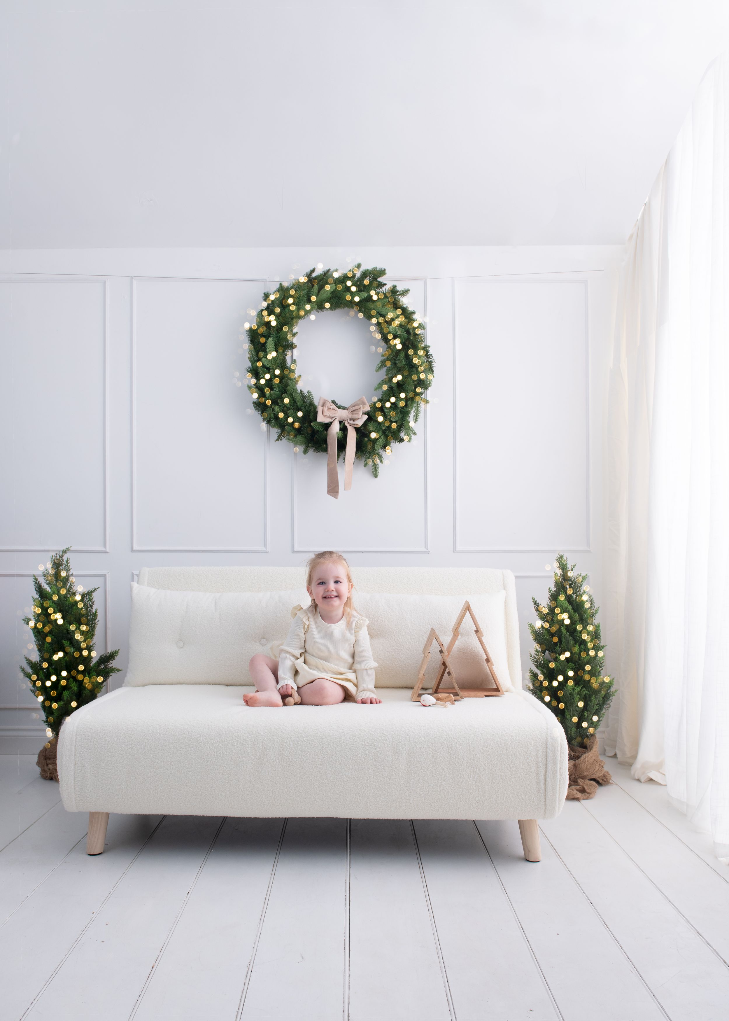 Baby in white onesie lying on white modern bed with holiday wreaths and decor in minimalist room.