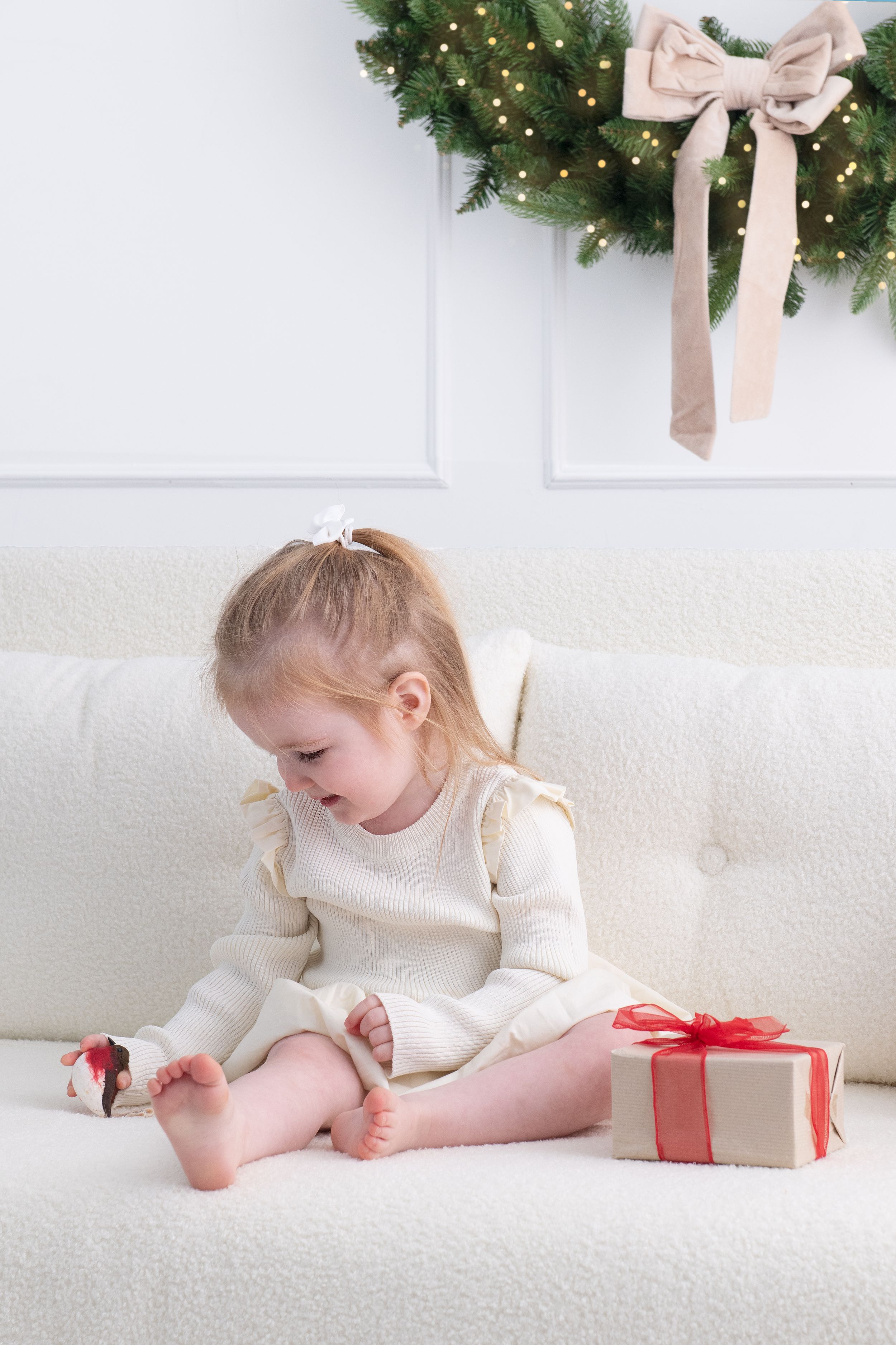 A small child in white sits on a white carpet near wrapped Christmas presents and holiday decorations.