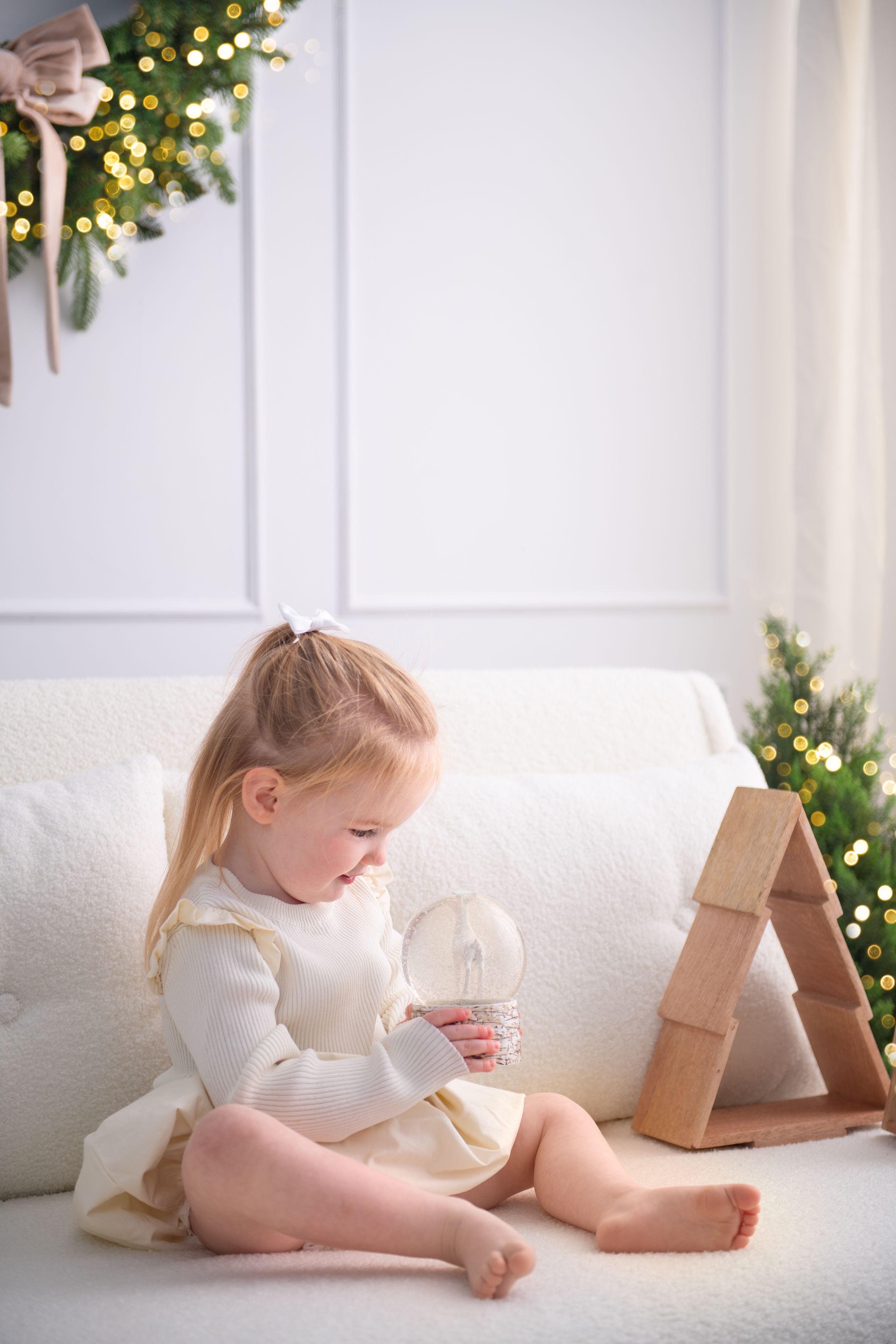A child in white reads a book while lying on a white couch with a Christmas tree twinkling in the background.